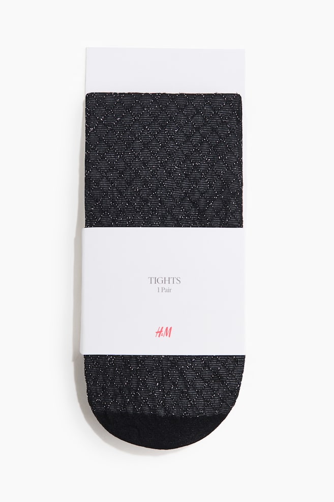 A pair of black tights features a textured diamond-knit pattern with subtle silver glitter flecks and a solid black toe. The tights are packaged flat against a white cardboard backing with a white paper band wrapped around the centre. - 1