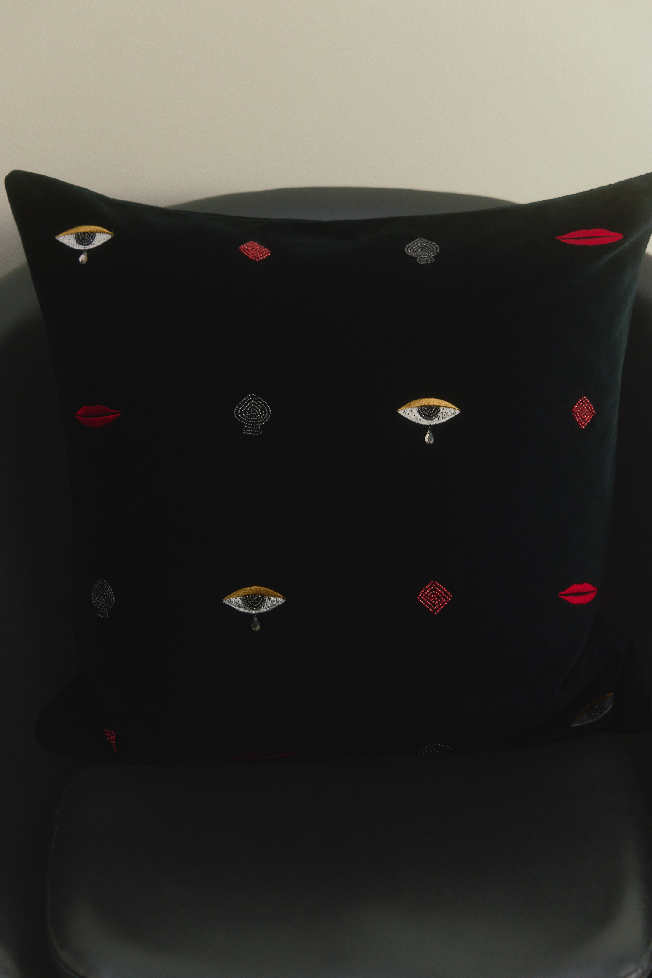 View larger image: A square black cotton velvet cushion cover is shown from the front, featuring a repeating grid of embroidered and beaded motifs. These decorative details depict crying eyes with shimmering stitching and dangling teardrop beads, red lips, silver spades, and red diamonds.