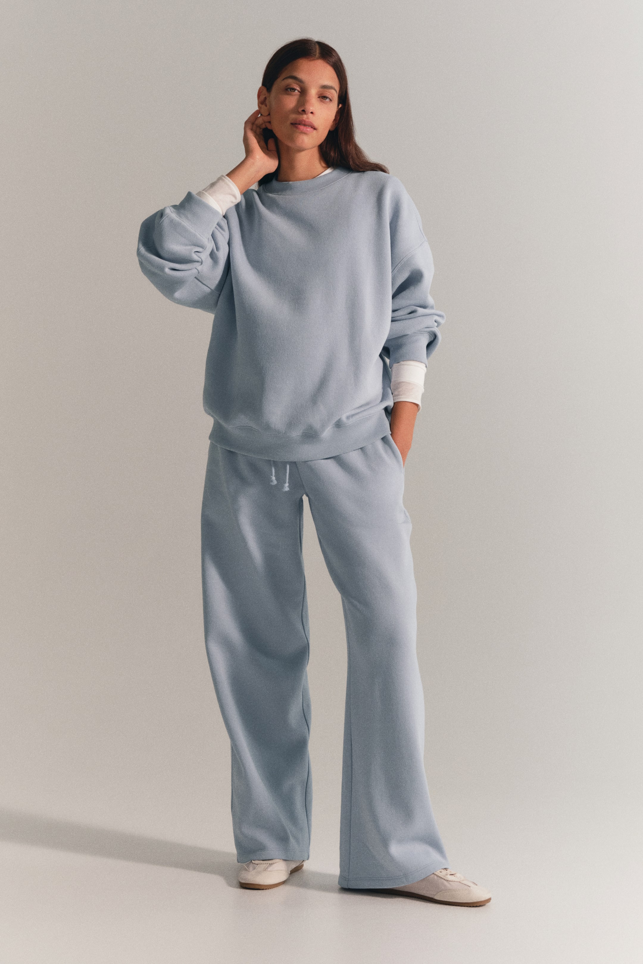 View larger image: The model is wearing a light blue sweat suit set, consisting of a crewneck sweater and matching wide-leg pants. The sweater features dropped shoulders and is layered over a white long-sleeve top, while the pants have an elastic drawstring waist and are styled with one hand in the pocket.