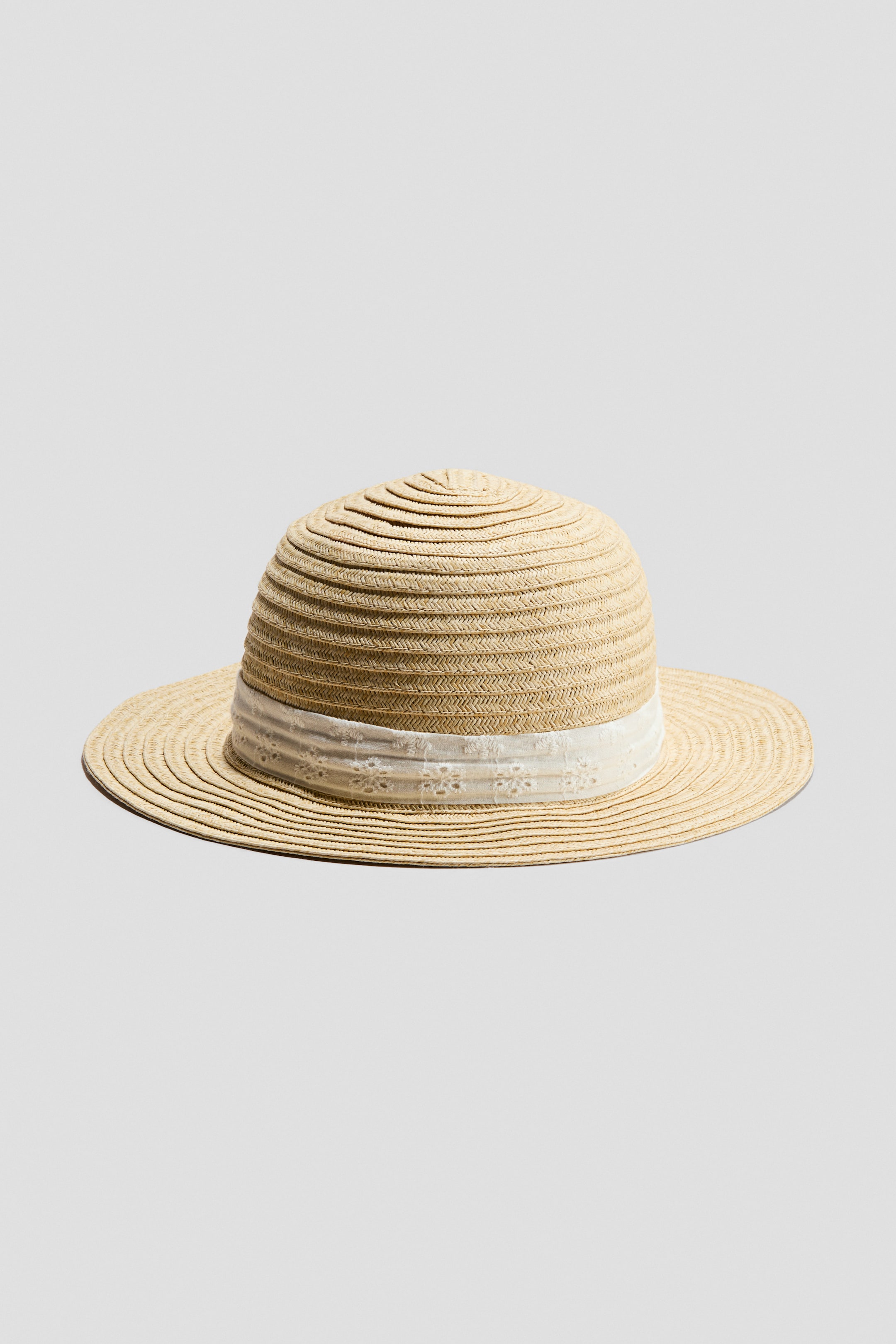Ampliar la imagen: Un sombrero de paja tejida presenta una corona redondeada y un ala ancha y plana. Una cinta blanca con un patrón floral de ojales rodea la base de la corona.