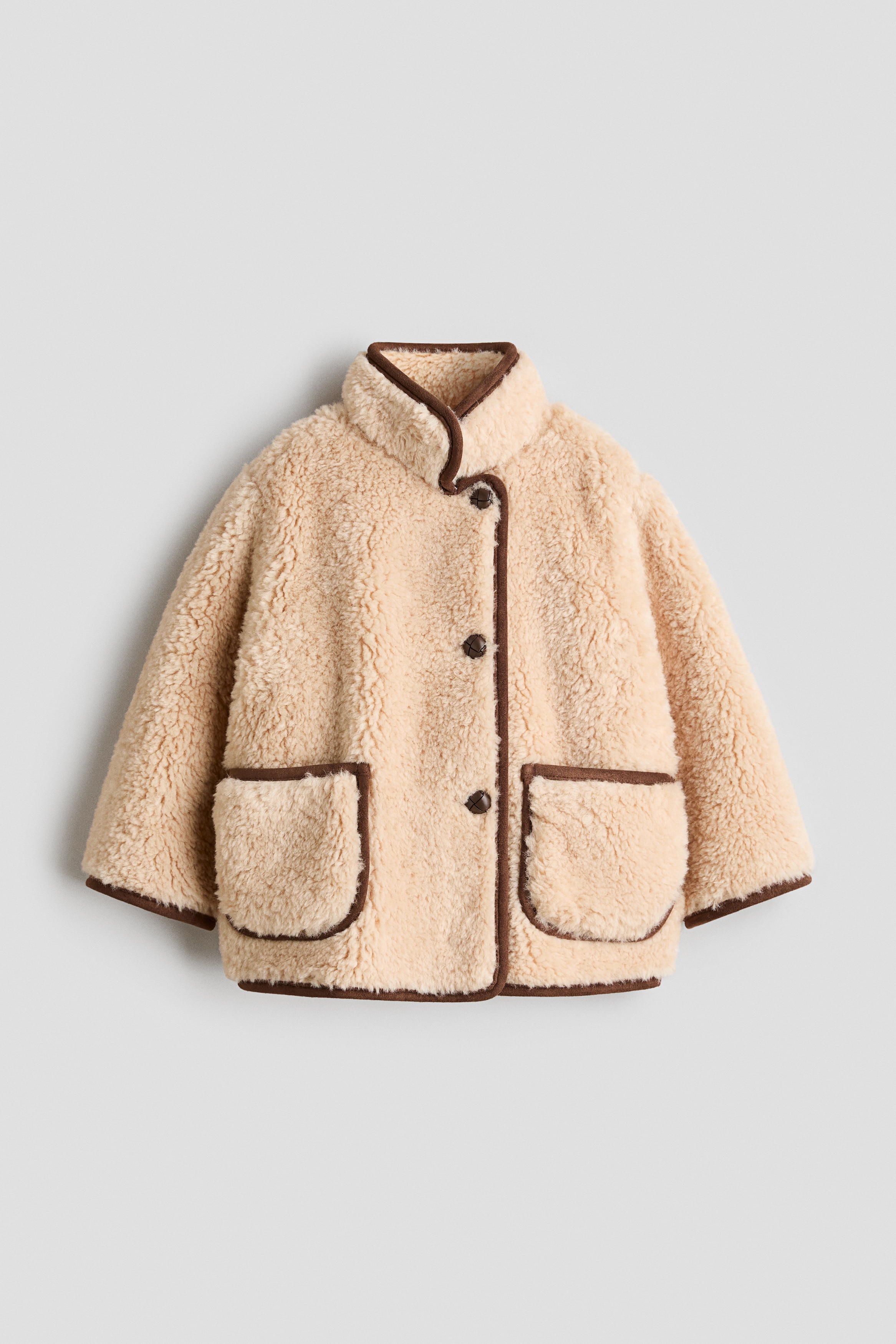 Una chaqueta de forro polar sherpa beige estÃ¡ orientada hacia el frente, con cuello alto, cierre asimÃ©trico de botones y ribete marrÃ³n oscuro en todos los bordes. La chaqueta presenta dos bolsillos de parche delanteros y mangas largas, ambos detallados con un ribete marrÃ³n a juego.