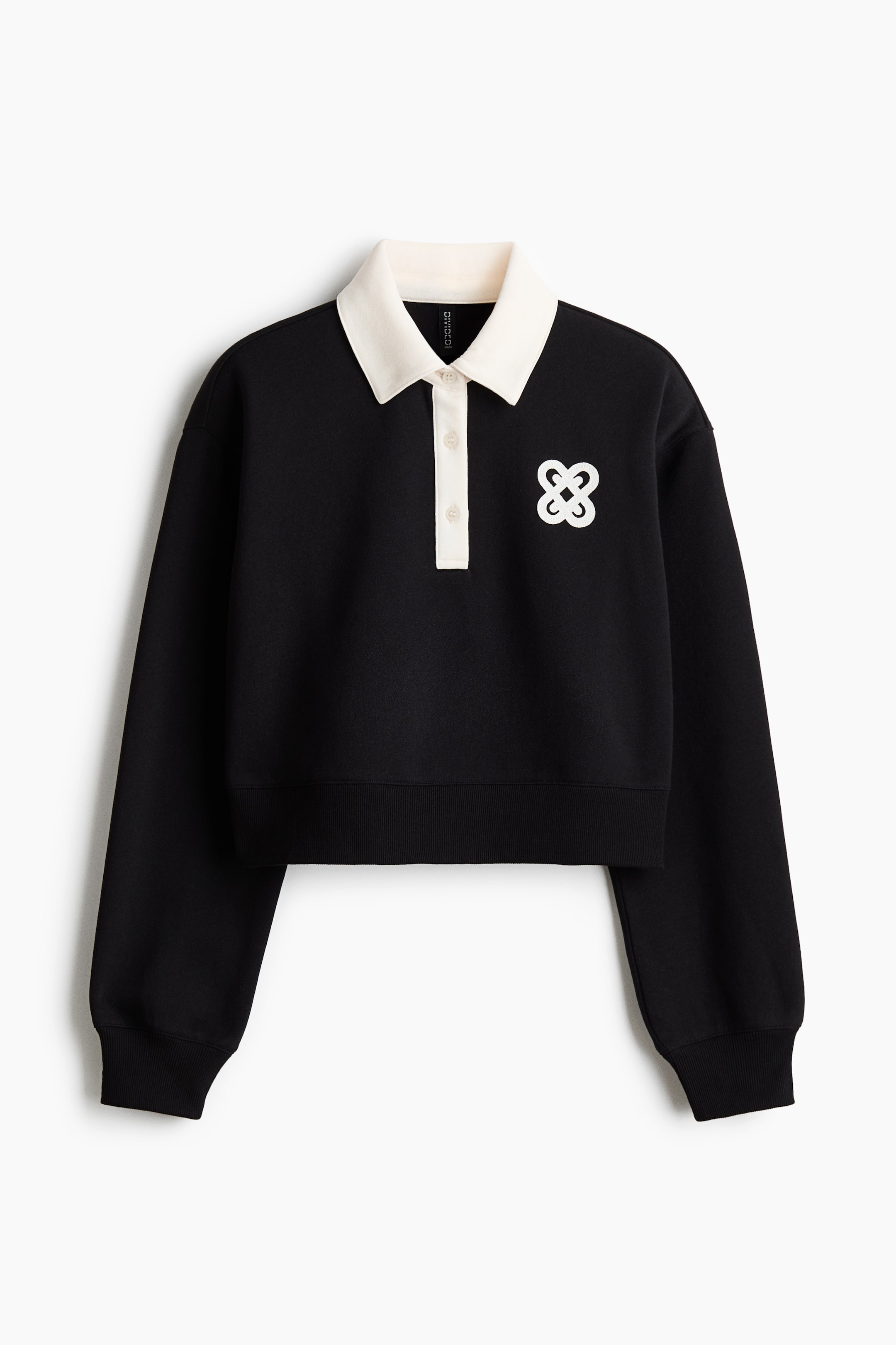 Xem ảnh lớn hơn: Một chiếc áo polo tay dài dáng ngắn màu đen đang hướng về phía TRƯỚC, thể hiện cổ áo dáng bèo màu kem và hàng nút ba cái. Một biểu tượng cách điệu màu trắng được trang trí ở ngực trái, và áo có cổ tay và gấu áo gân.