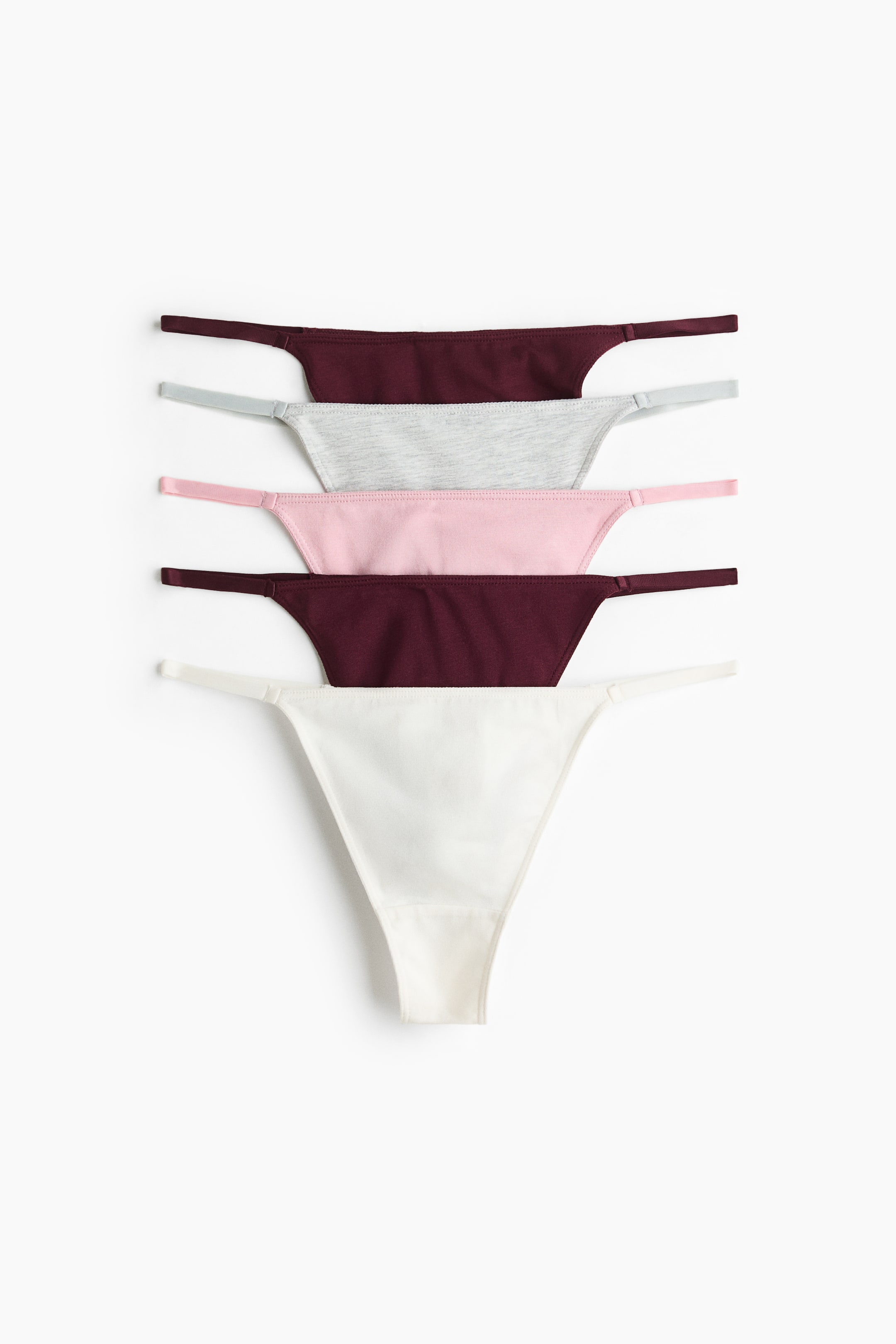 Grotere afbeelding bekijken: Vijf G-stringonderbroeken liggen plat in een verticale stapel met de voorkant naar voren, met een lage taille en smalle elastische zijbandjes. De set toont van boven naar beneden de kleuren donker bordeaux, gemêleerd grijs, lichtroze, donker bordeaux en gebroken wit, waarbij het onderste paar volledig het driehoekige voorpaneel en het gevoerde kruis laat zien.