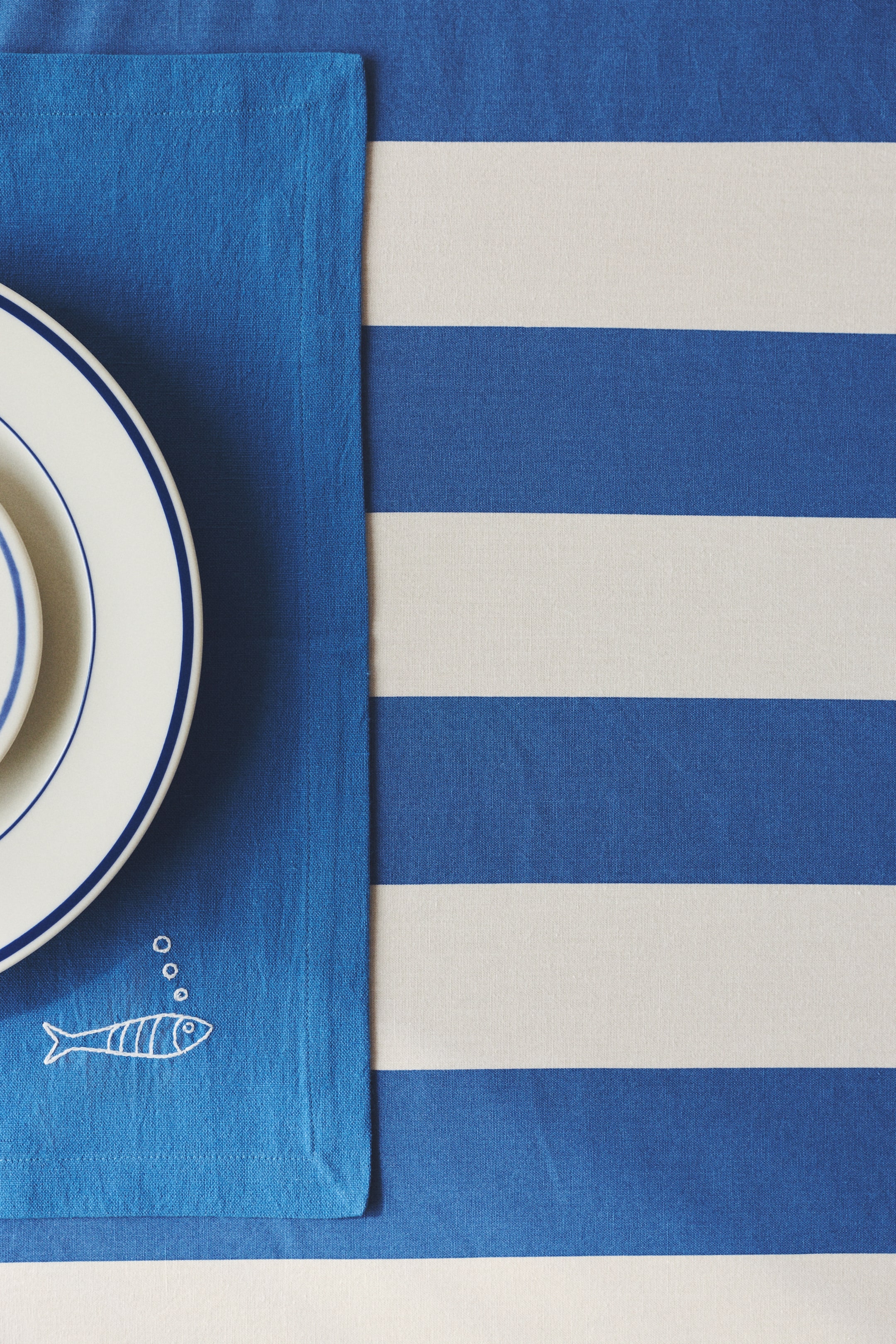 View larger image: A blue placemat is laid out flat, showcasing a white embroidered fish motif with three bubbles in the bottom left corner. A stack of white plates with blue rims sits atop the placemat, partially obscuring a blue and white striped tablecloth.