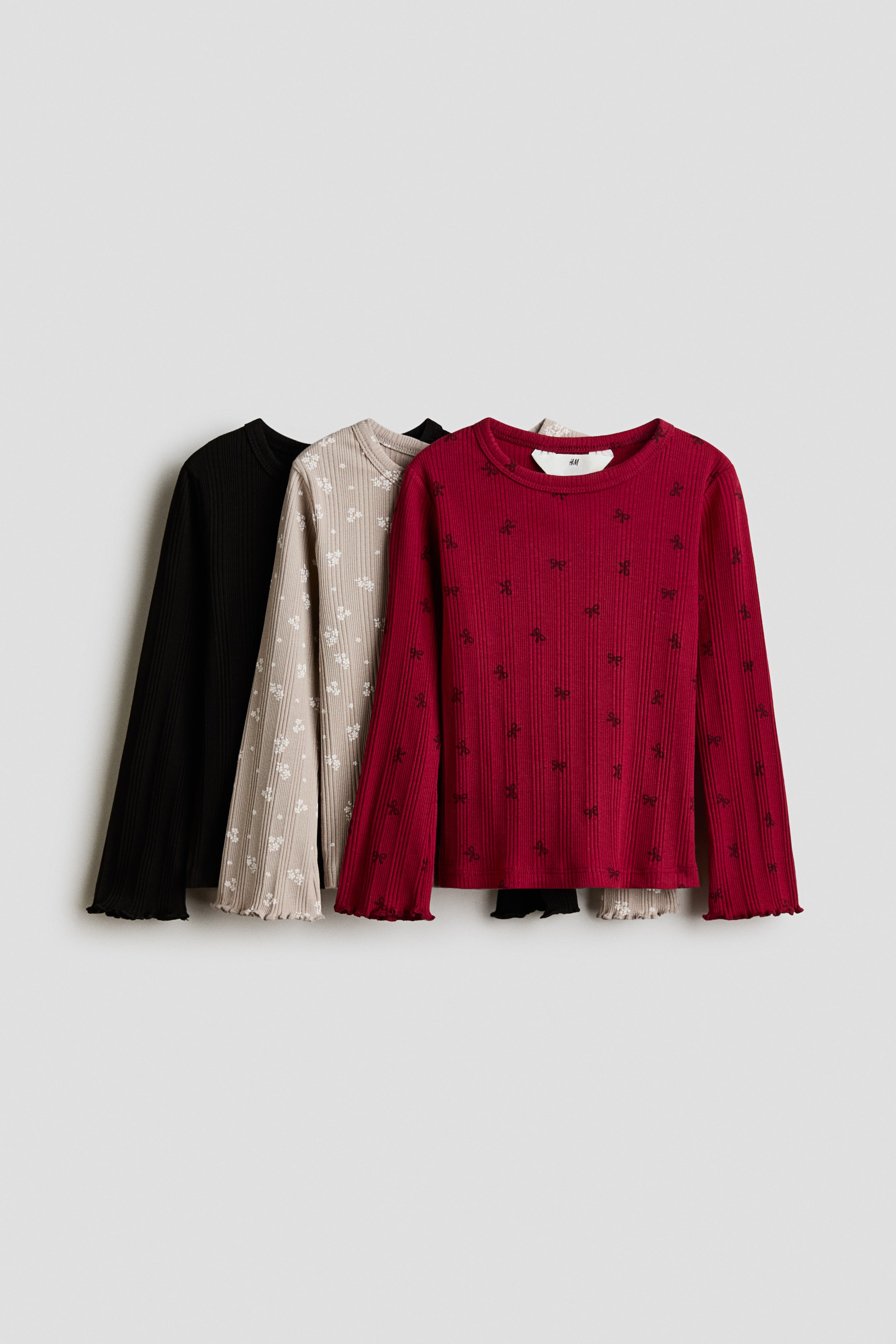 Größeres Bild anzeigen: Drei langärmelige Tops aus gerippter Baumwolljersey sind in einer Lagenanordnung von vorne zu sehen. Das Set umfasst ein dunkelrotes Top mit kleinem schwarzen Schleifenmuster, ein hellbeiges Top mit weißem Blumenprint sowie ein einfarbig schwarzes Top. Jedes Modell verfügt über einen Rundhalsausschnitt und lange Ärmel mit Overlock-Abschlüssen an den Bündchen.