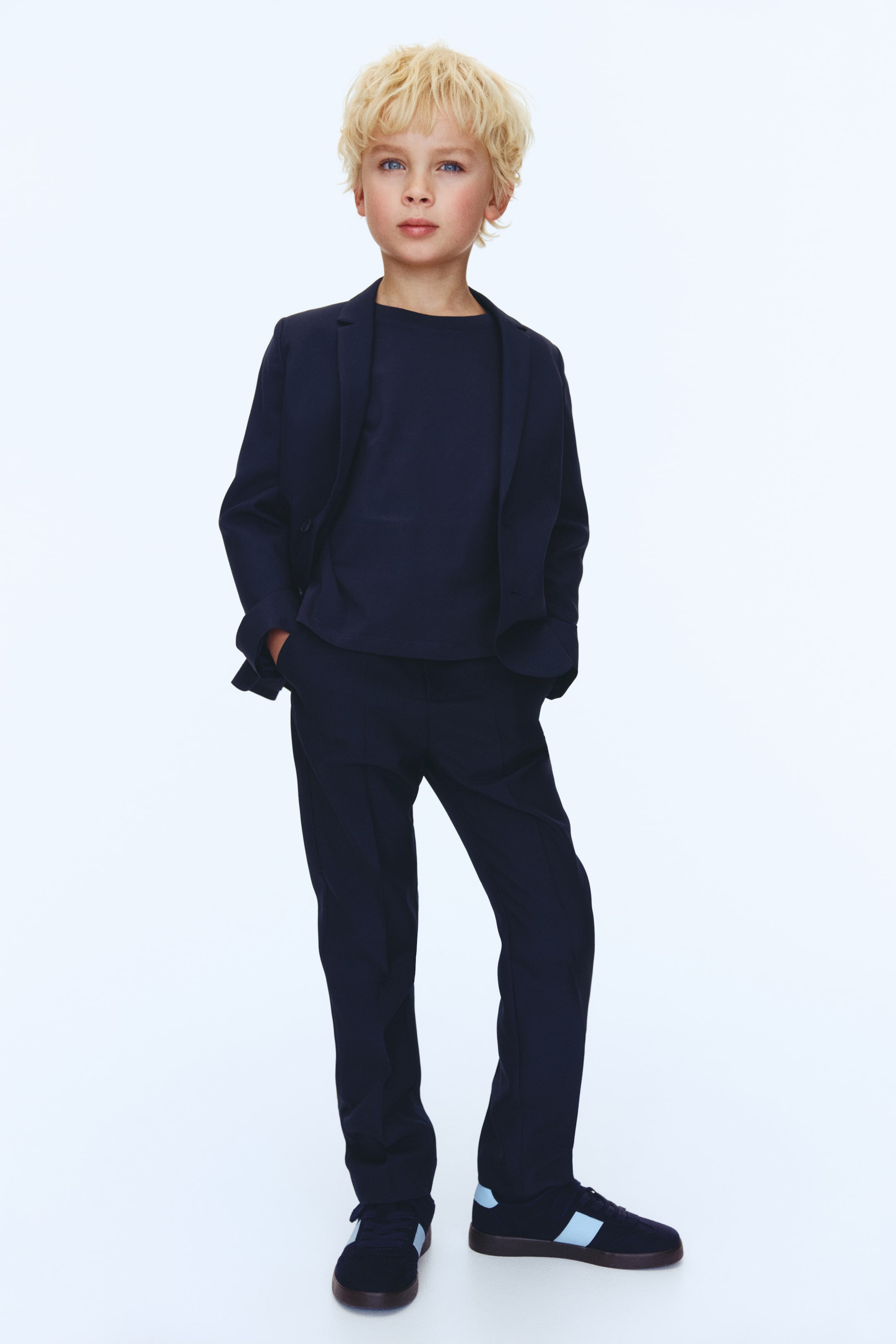 Zobacz większy obraz: Granatowa marynarka noszona jest otwarta na granatowym t-shircie z okrągłym dekoltem, w połączeniu z granatowymi spodniami o prostym kroju z przednimi kantami. Granatowe niskie trampki z jasnoniebieskimi paskami po bokach dopełniają stylizację.