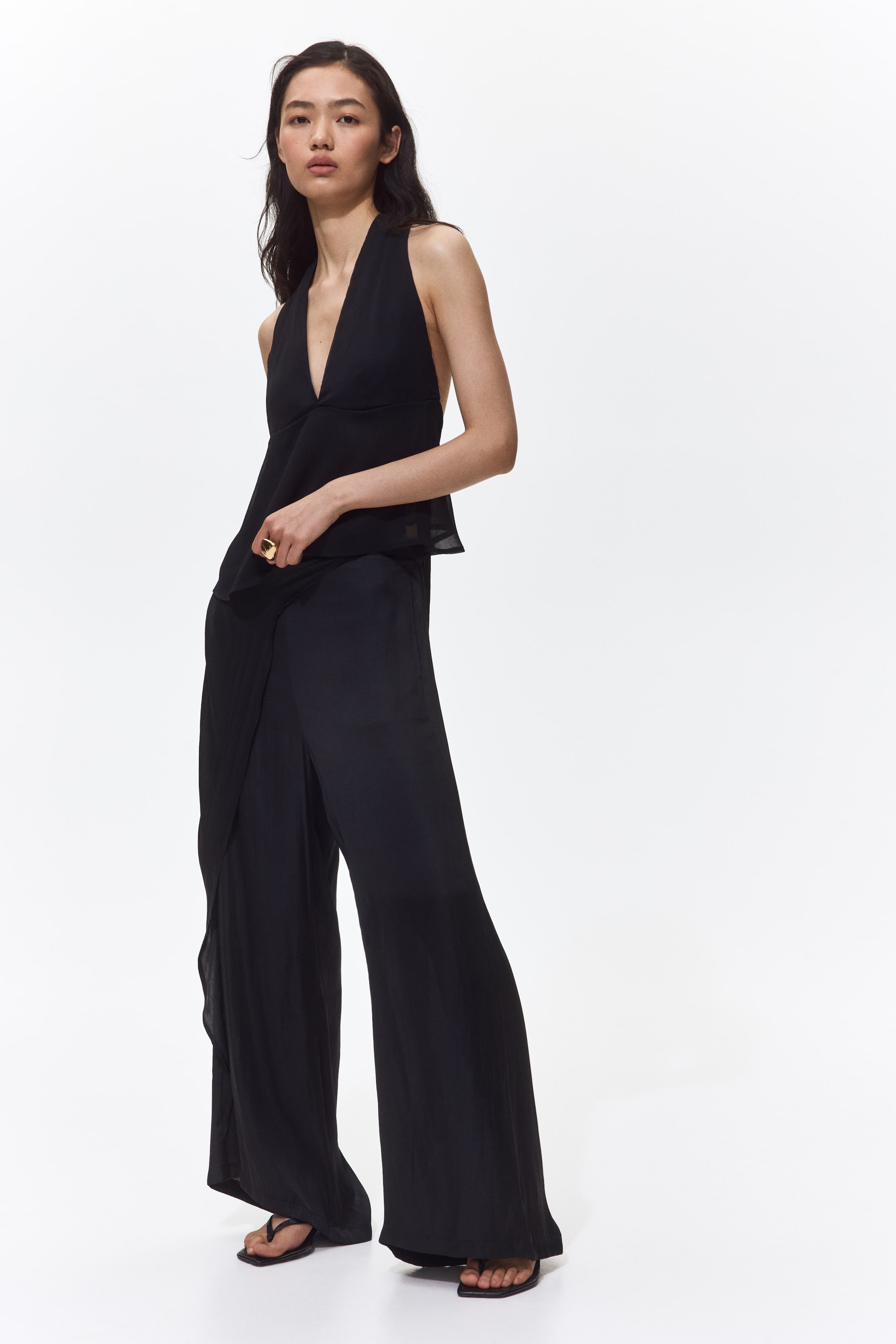 Ampliar la imagen: Un conjunto coordinado compuesto por un top halter negro y pantalones de pierna ancha negros. El top halter presenta un escote en V profundo y un dobladillo fluido con capas transparentes, mientras que los pantalones de pierna ancha exhiben un diseño de frente cruzado en una pierna y un ajuste relajado de largo completo.