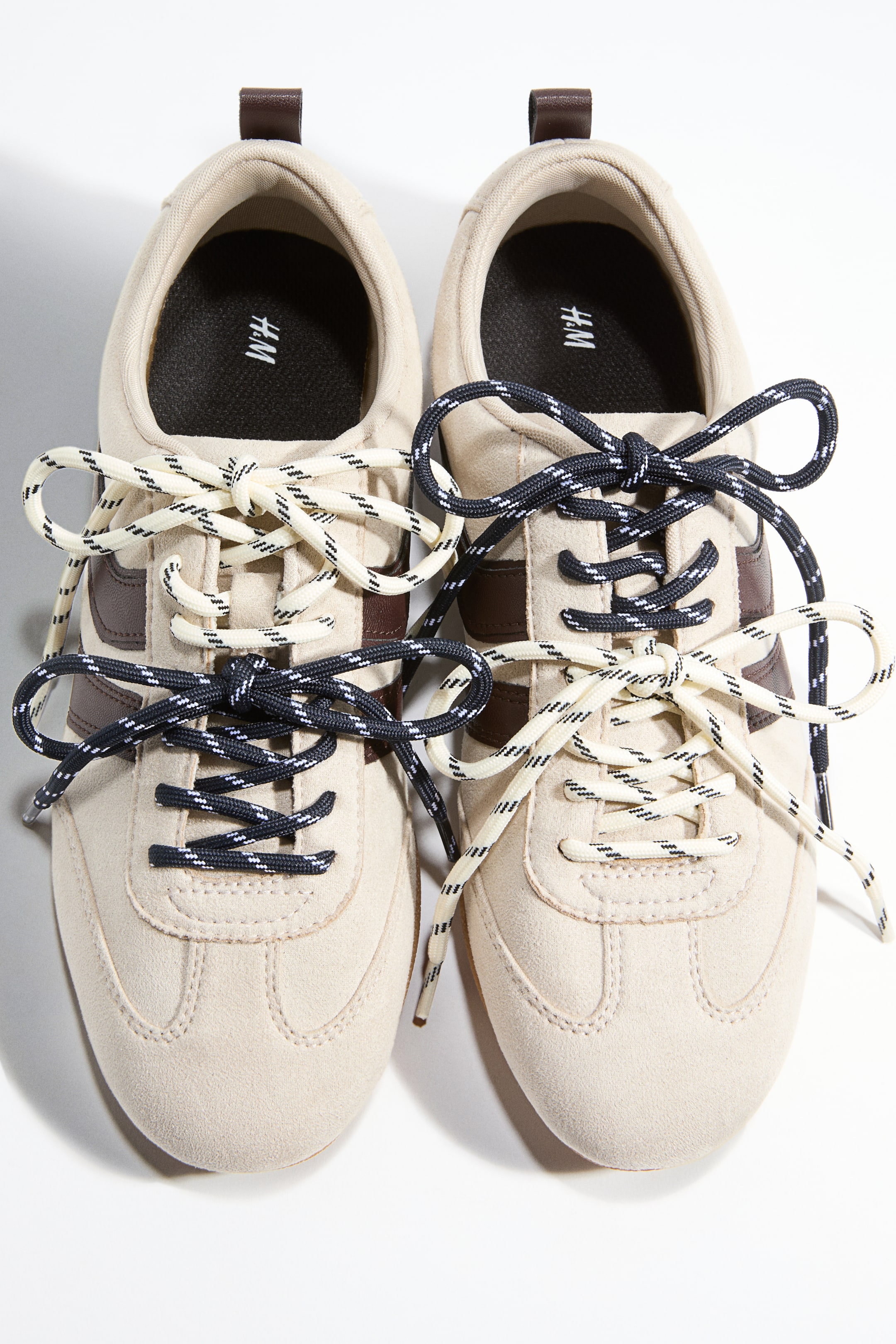 View larger image: Two sets of patterned shoelaces are tied on beige trainers, viewed from directly above. One set is cream with black stripes, and the other is navy blue with white stripes, both forming bows.