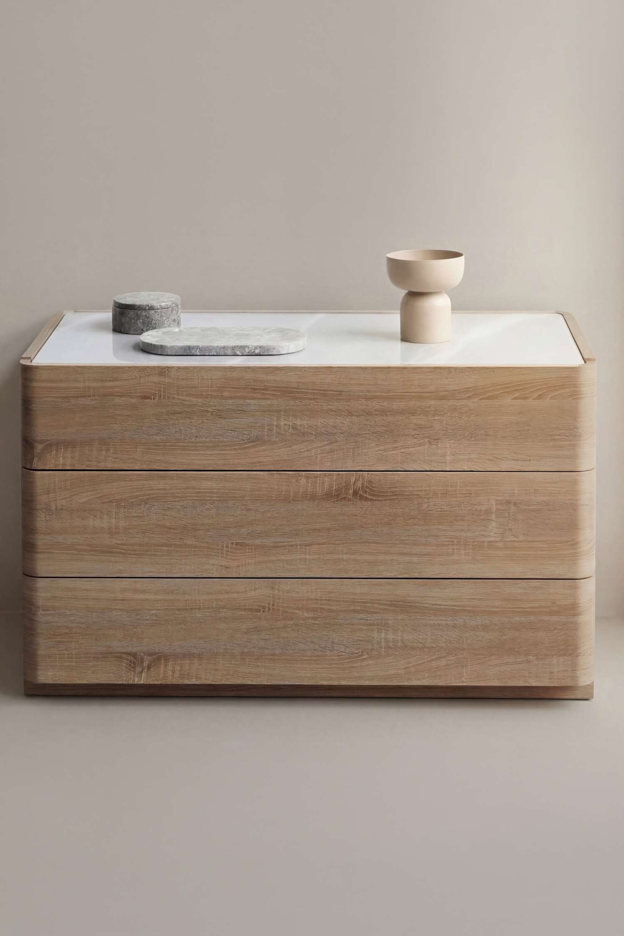 Visualizza immagine più grande: Una cassettiera in legno chiaro a tre cassetti presenta un piano superiore bianco lucido e bordi arrotondati. Sopra sono posati oggetti decorativi in pietra grigia e un vaso scultoreo beige.