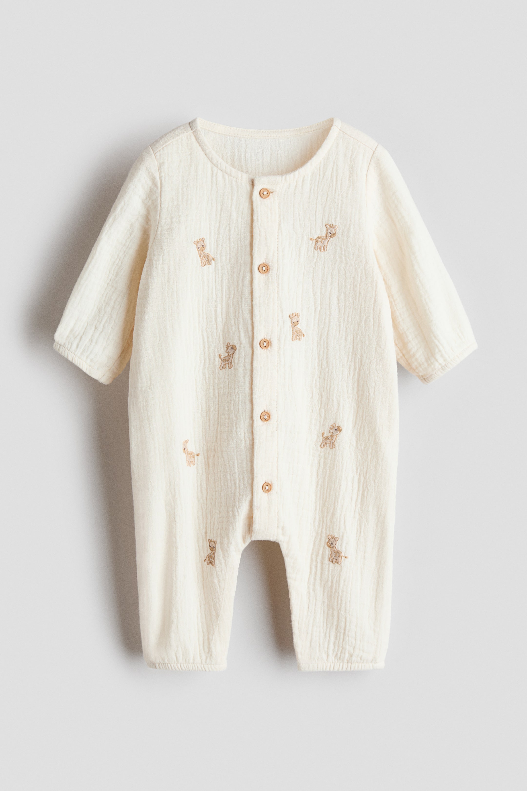 View larger image: A cream-colored baby romper is presenting FRONT, featuring long sleeves, a round neck, and a full-length front placket with wooden buttons, and is adorned with scattered light brown giraffe embroidery across its crinkled fabric.