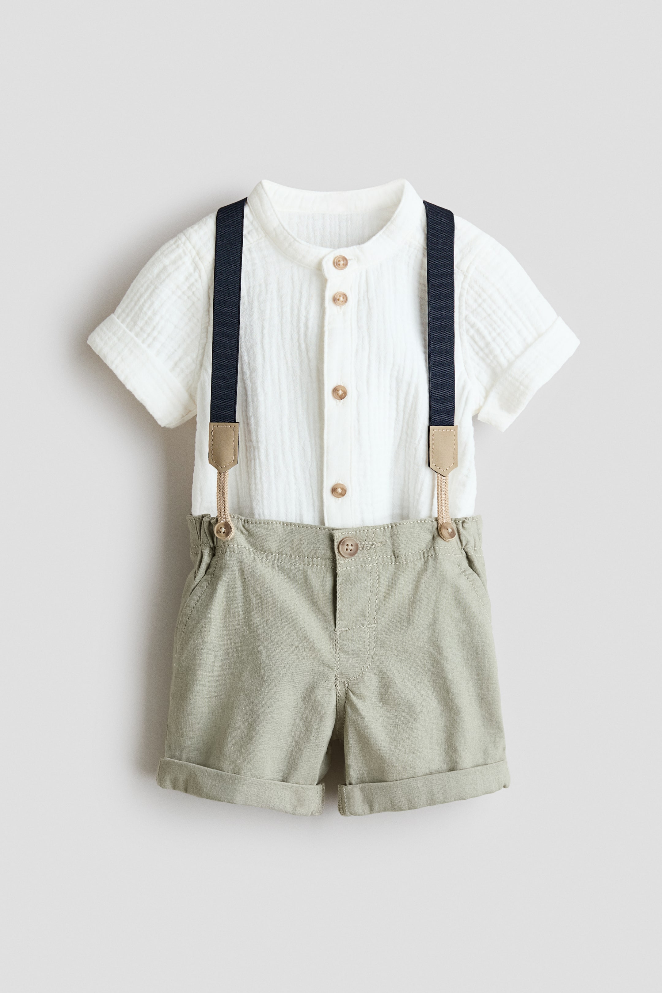 View larger image: Front view of a white textured short-sleeved shirt and light-green shorts set. The shirt features a band collar, cuffed sleeves and light-brown buttons down the front. The shorts feature dark-blue elasticated braces with tan leather-look tabs, an adjustable elasticated waist with a mock fly and button, diagonal mock pockets and rolled cuffs.