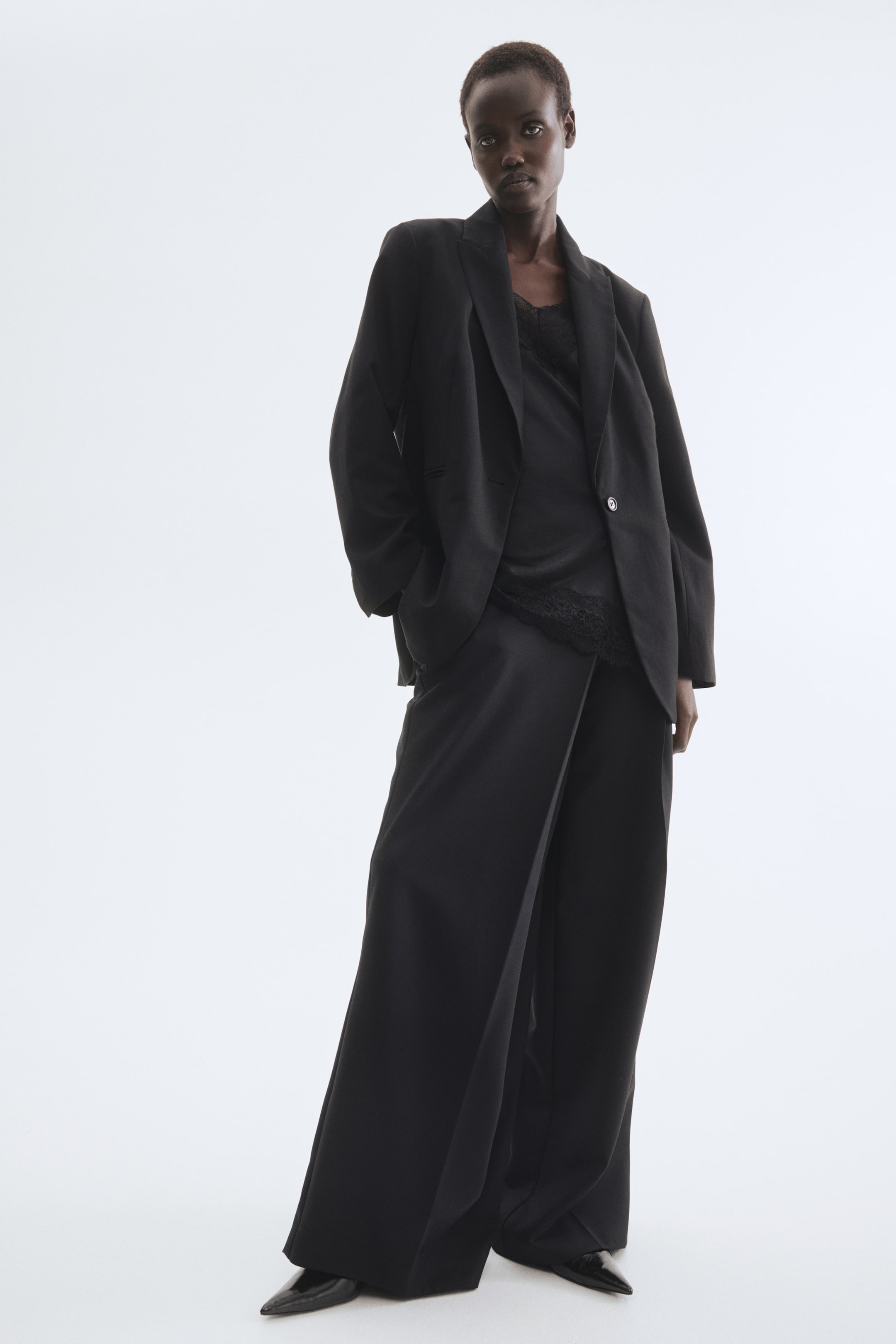 Größeres Bild ansehen: Ein schwarzer einreihiger Blazer mit Spitzfasson, strukturierten Schultern, langen Ärmeln, einem vorderen Knopf und paspelierten Vordertaschen wird von dem Modell offen über einem schwarzen Camisole getragen. Das Modell lässt ihre Hände in den Taschen des Blazers ruhen.