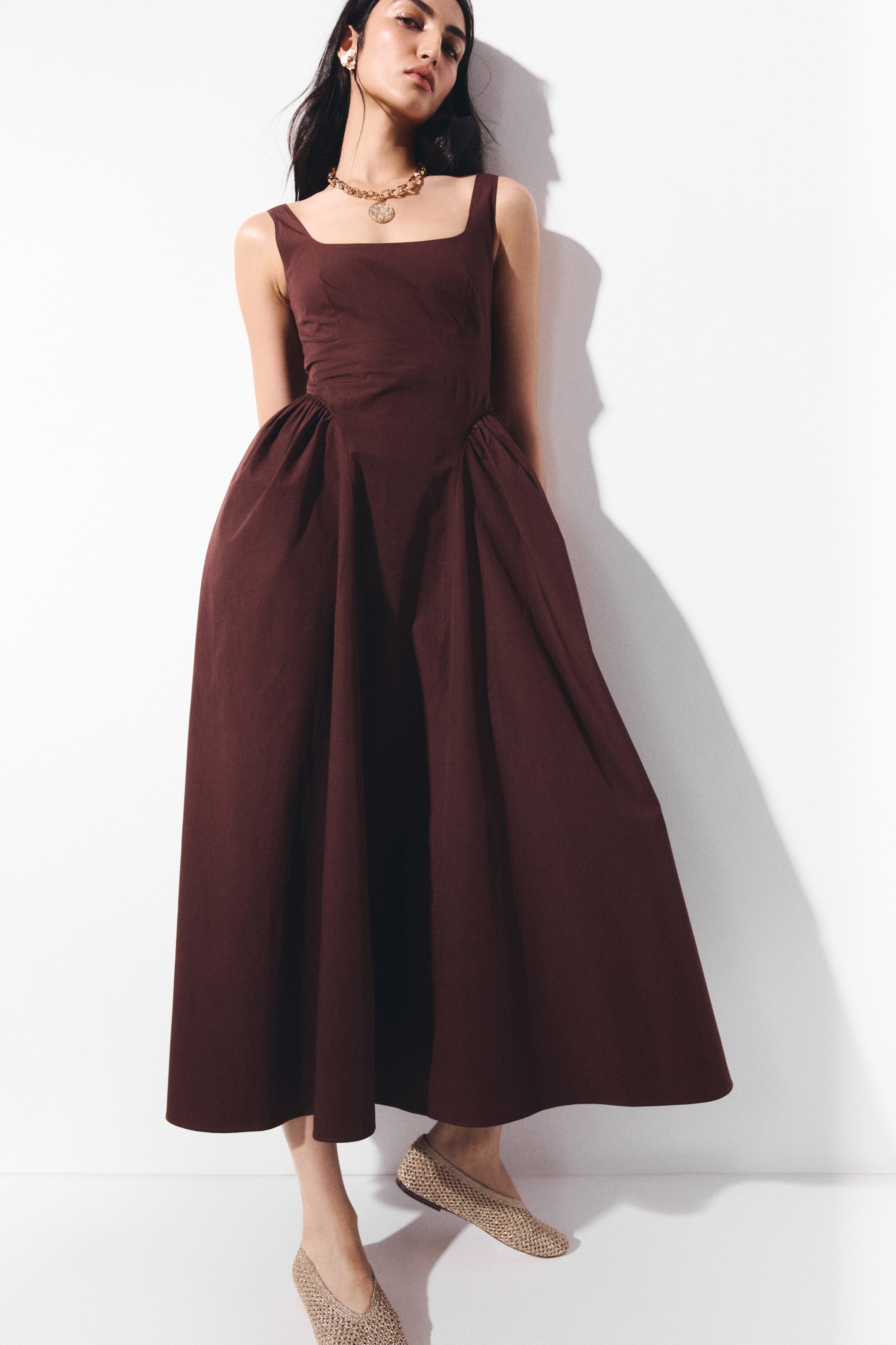 View larger image: The dark brown midi dress showcases a square neckline with broad straps. Its fitted bodice flows into a full skirt, which gathers prominently at the side seams.