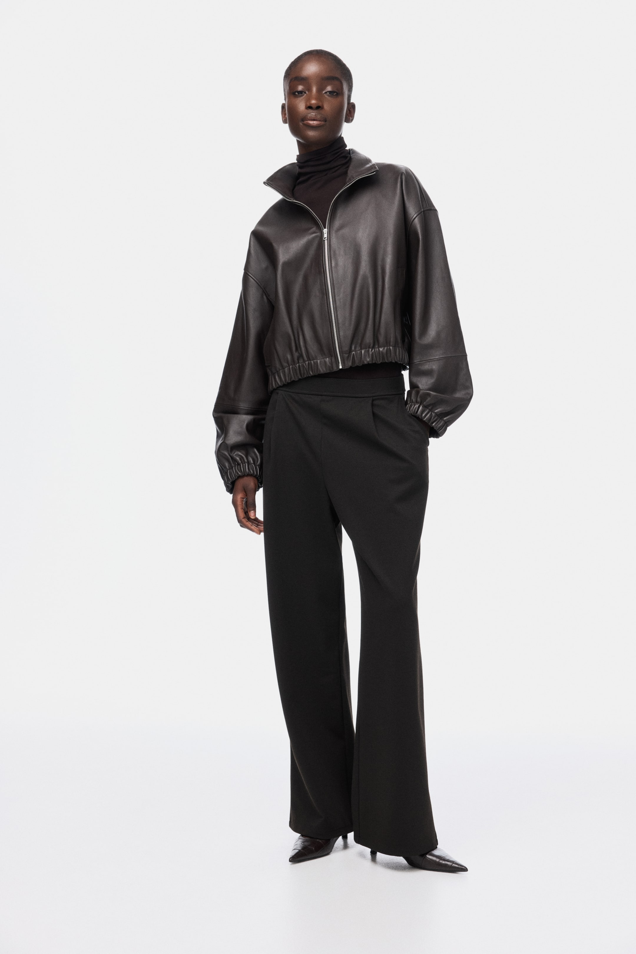 View larger image: A dark brown leather bomber jacket is being worn, featuring a full-length silver zipper, a stand-up collar, and elasticized hem and cuffs that create a cropped, voluminous silhouette. It is layered over a dark turtleneck.