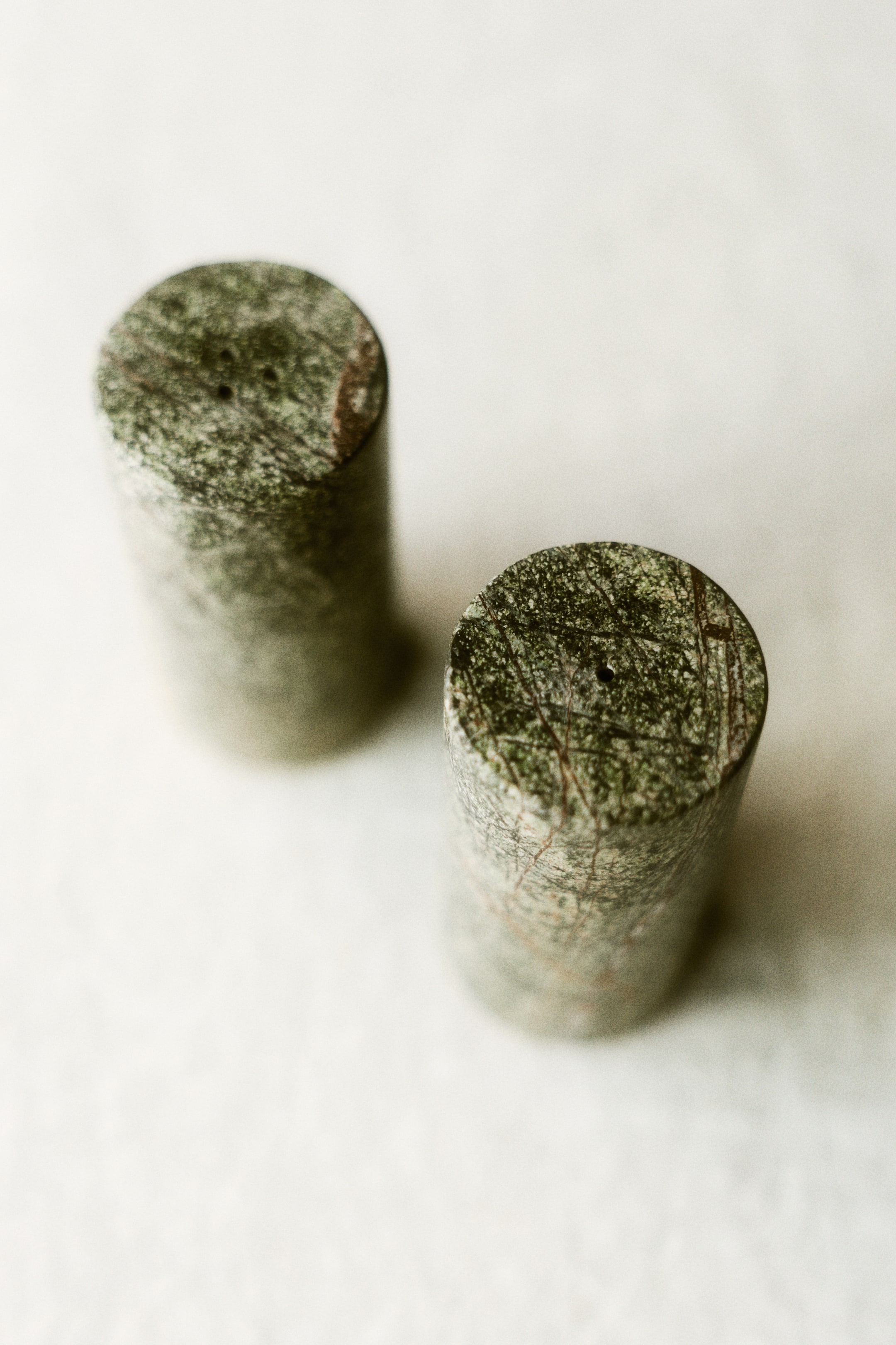 View larger image: Two cylindrical objects crafted from mottled green and gray stone with reddish-brown veining stand upright. The front object showcases a small, dark hole on its flat top surface, while the second object is positioned slightly behind and to its left.