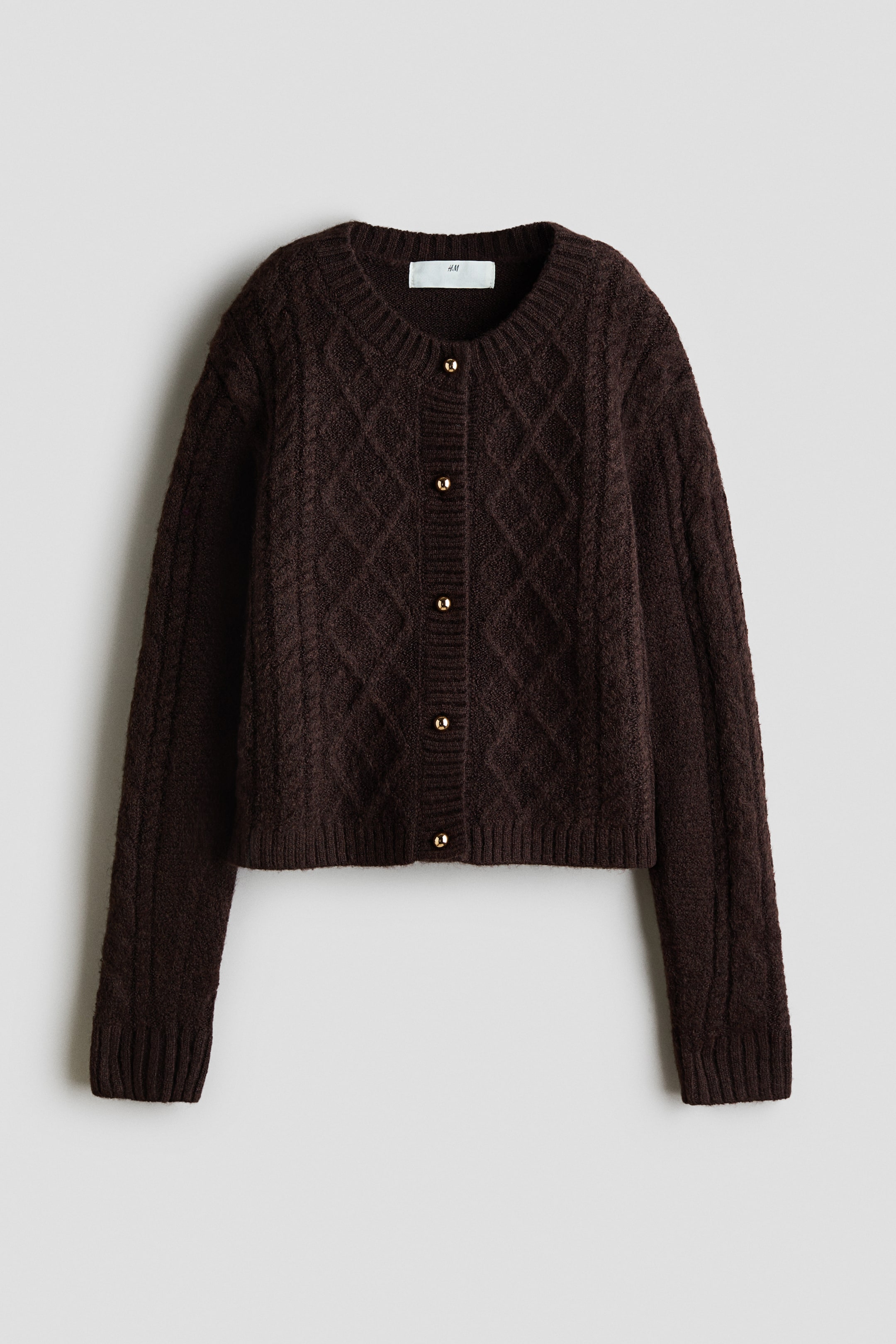 Ver imagen más grande: Un cárdigan marrón oscuro muestra su parte frontal, con una suave textura de punto trenzado. Presenta un cuello redondo rematado en canalé, botones de color metalizado a lo largo de la tapeta abotonada acanalada y puños y bajo acanalados.