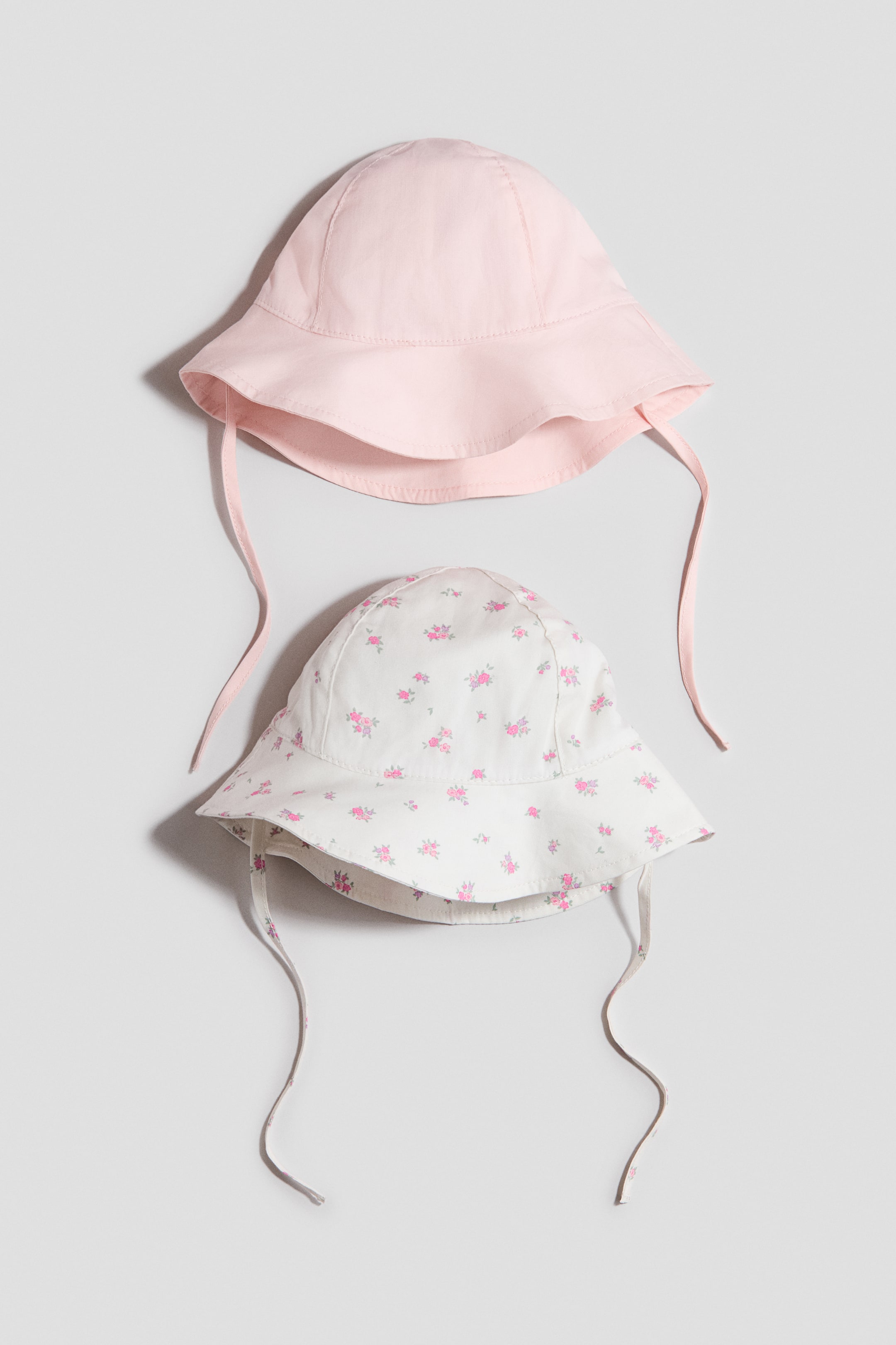 Ver imagen más grande: Sombreros de sol en un tejido de algodón aireado con lazos bajo la barbilla se exhiben desde el frente. El sombrero superior presenta un color rosa claro sólido, y el sombrero inferior presenta un fondo blanco con un pequeño estampado floral rosa.