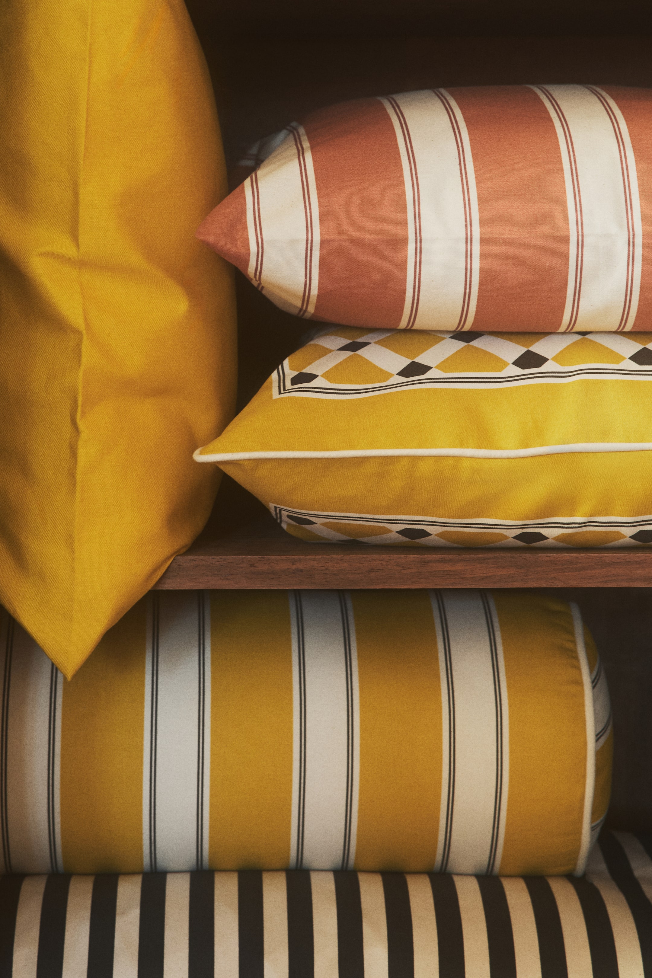 View larger image: Presenting a curated collection of cushions, prominently featuring a large, solid yellow cushion standing tall. Arranged horizontally are a rectangular cushion adorned with terracotta and white vertical stripes, a yellow rectangular cushion accented with white piping and a geometric diamond border, a cylindrical bolster cushion displaying yellow and white vertical stripes, and a dark striped cushion subtly peeking from beneath.