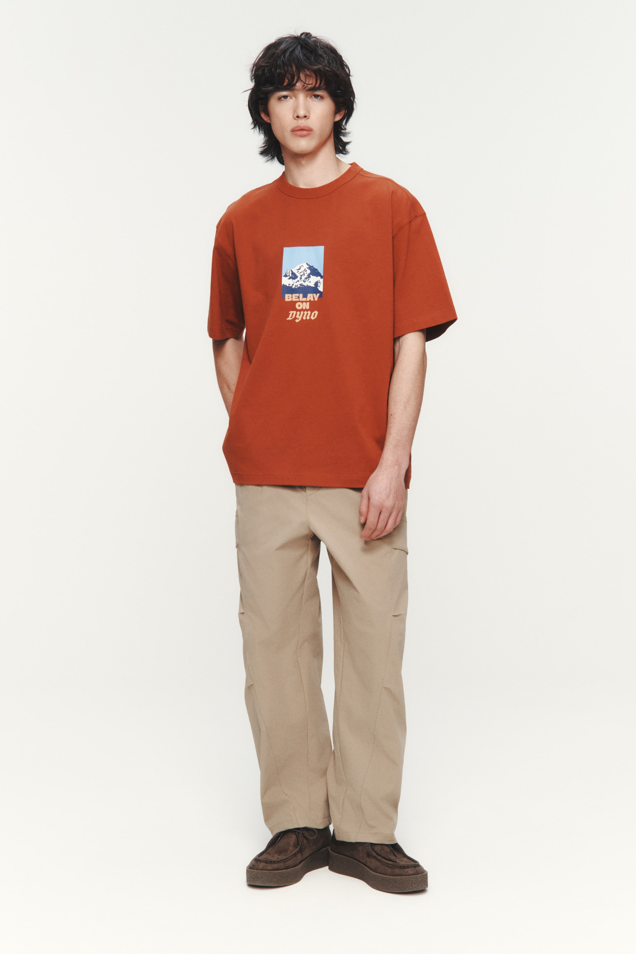 View larger image: The rust orange t-shirt showcases a relaxed fit and short sleeves, complete with a ribbed crew neck. On the front chest, a rectangular graphic illustrates snowy mountains beneath a blue sky, accompanied by the text "BELAY ON DYNO" below.