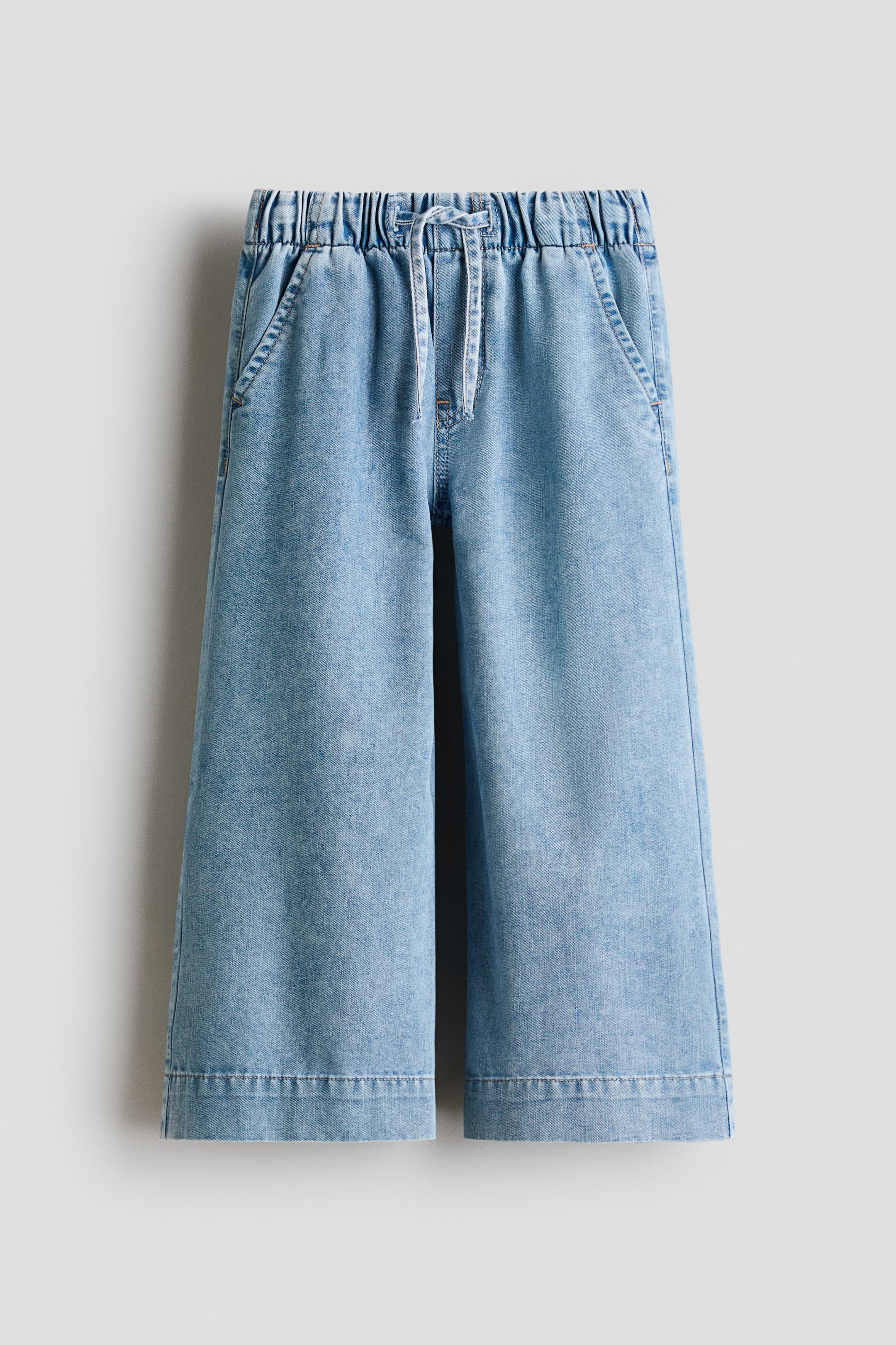 View larger image: Loose-fit, light blue denim trousers are showcased from the front, featuring wide legs. They are crafted with an elasticated, drawstring waist, a mock fly, and diagonal side pockets.