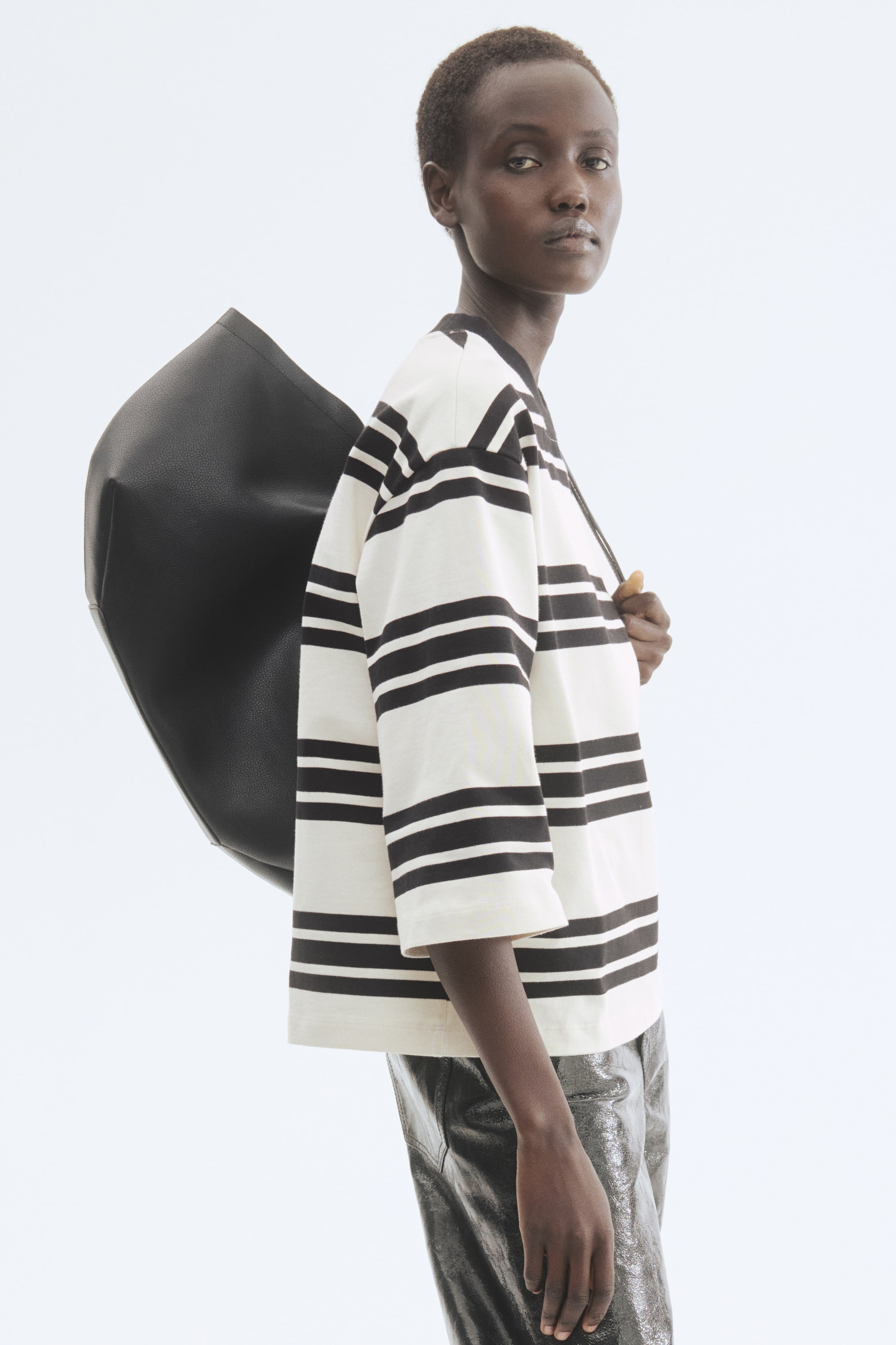 Agrandir l'image: Un haut ample avec de larges rayures horizontales noires et blanc cassé et des manches trois-quarts est présenté de profil. Un grand sac à bandoulière noir, non structuré, avec une texture granulée est porté, tombant de l'épaule dans le dos.