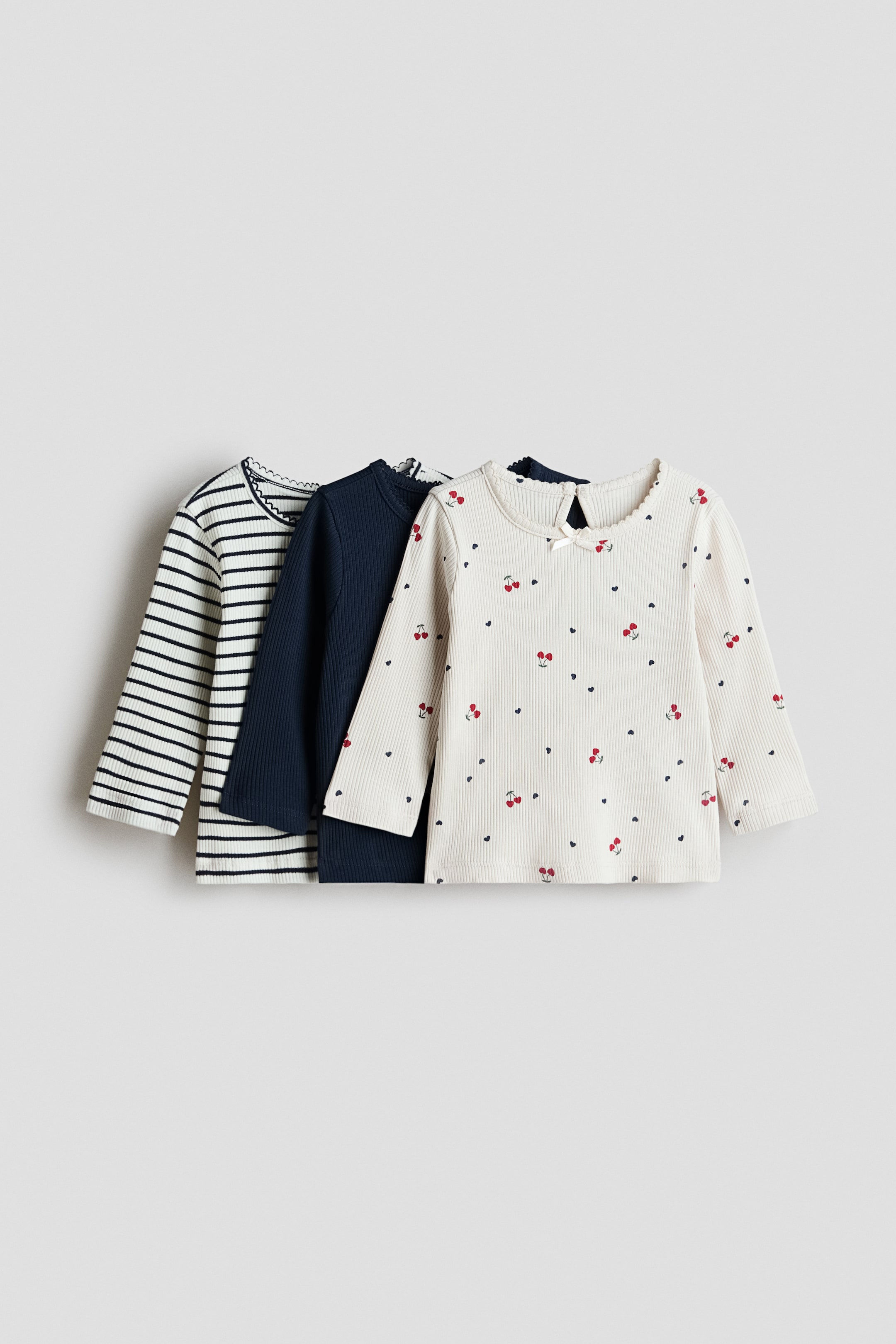 View larger image: Three long-sleeved ribbed tops are showcasing their front sides in a slightly overlapping arrangement. The leftmost top is white with navy horizontal stripes, the middle is solid navy blue, and the rightmost is off-white with a repeating cherry and heart pattern. All three tops are featuring scalloped necklines, and the off-white top also includes a small white bow at its visible back keyhole opening.