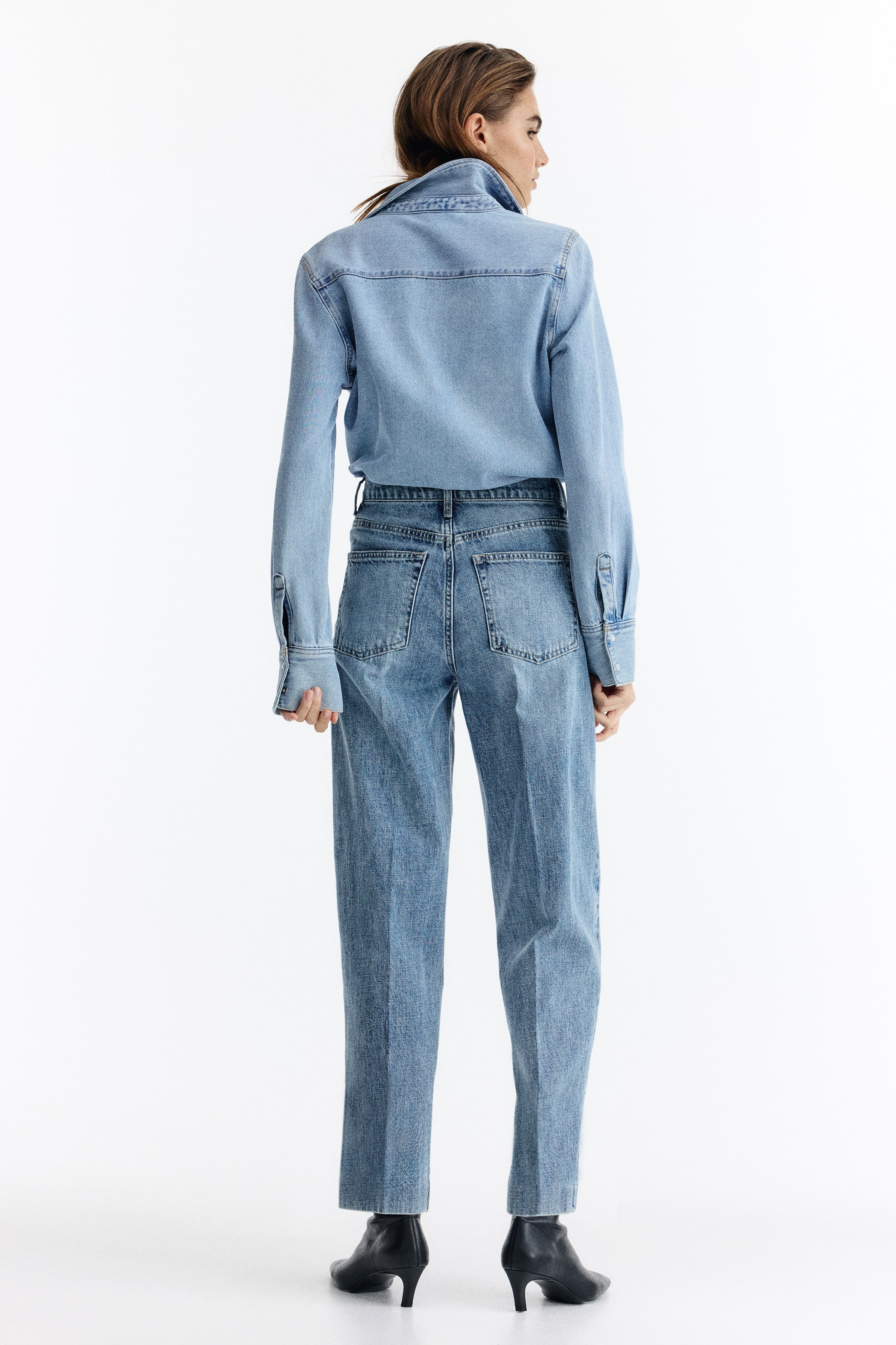 Grotere afbeelding bekijken: Een lichtblauw denim overhemd, met een rugpas en lange mouwen met knoopmanchetten, wordt van achteren getoond en is ingestopt in een hoog getailleerde, middelblauwe denim jeans. De jeans hebben twee achterzakken en een ontspannen pasvorm, en worden gedragen met zwarte enkelboots.