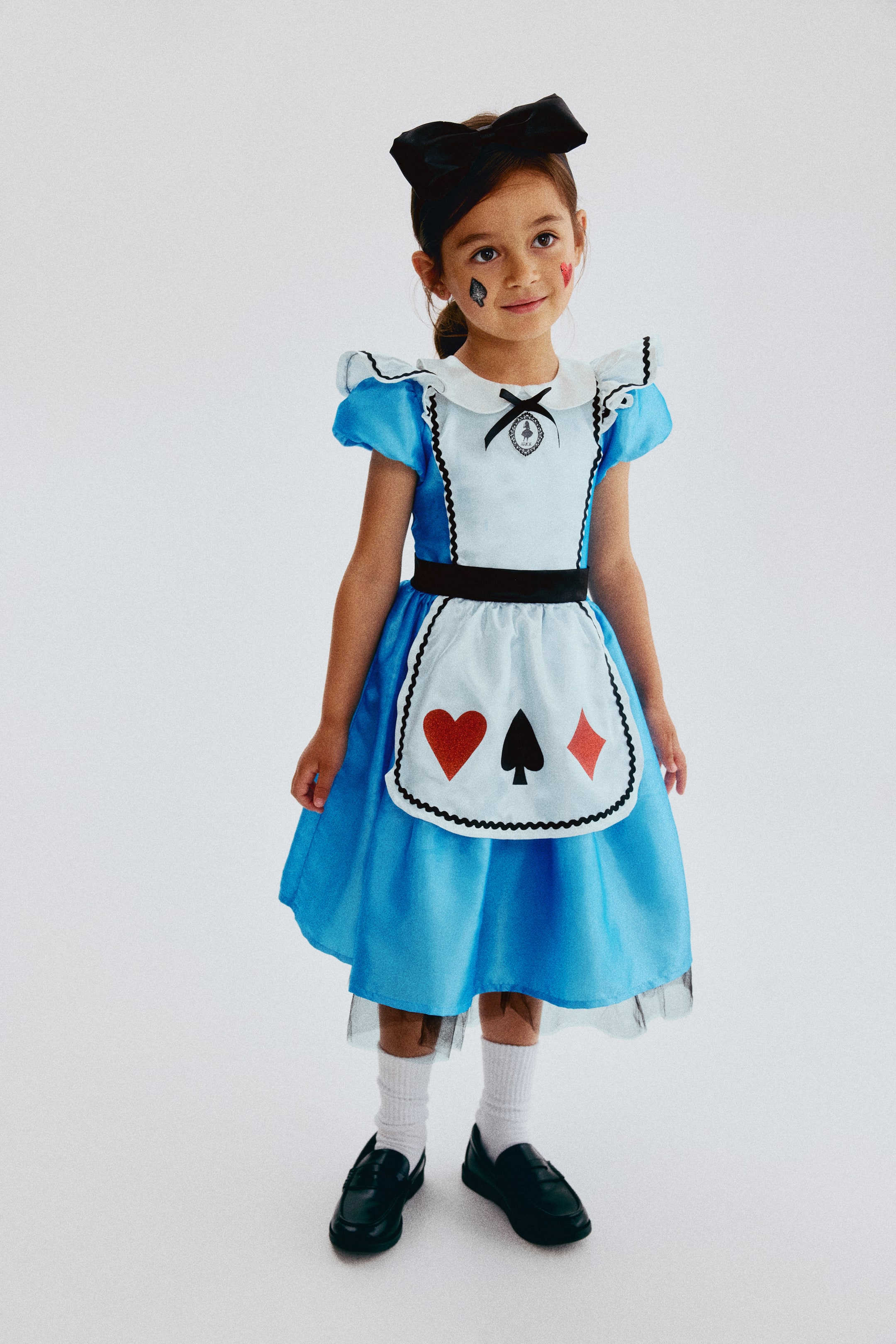View larger image: A child’s costume consists of a blue dress with short puff sleeves, a white Peter Pan collar, and black trim along the bodice and sleeves. A white apron, trimmed in black and featuring red heart, black spade, and red diamond card suit symbols, is layered over the dress with a black waistband, while a black tulle underskirt is visible beneath the blue skirt. A black bow headband tops off the ensemble.