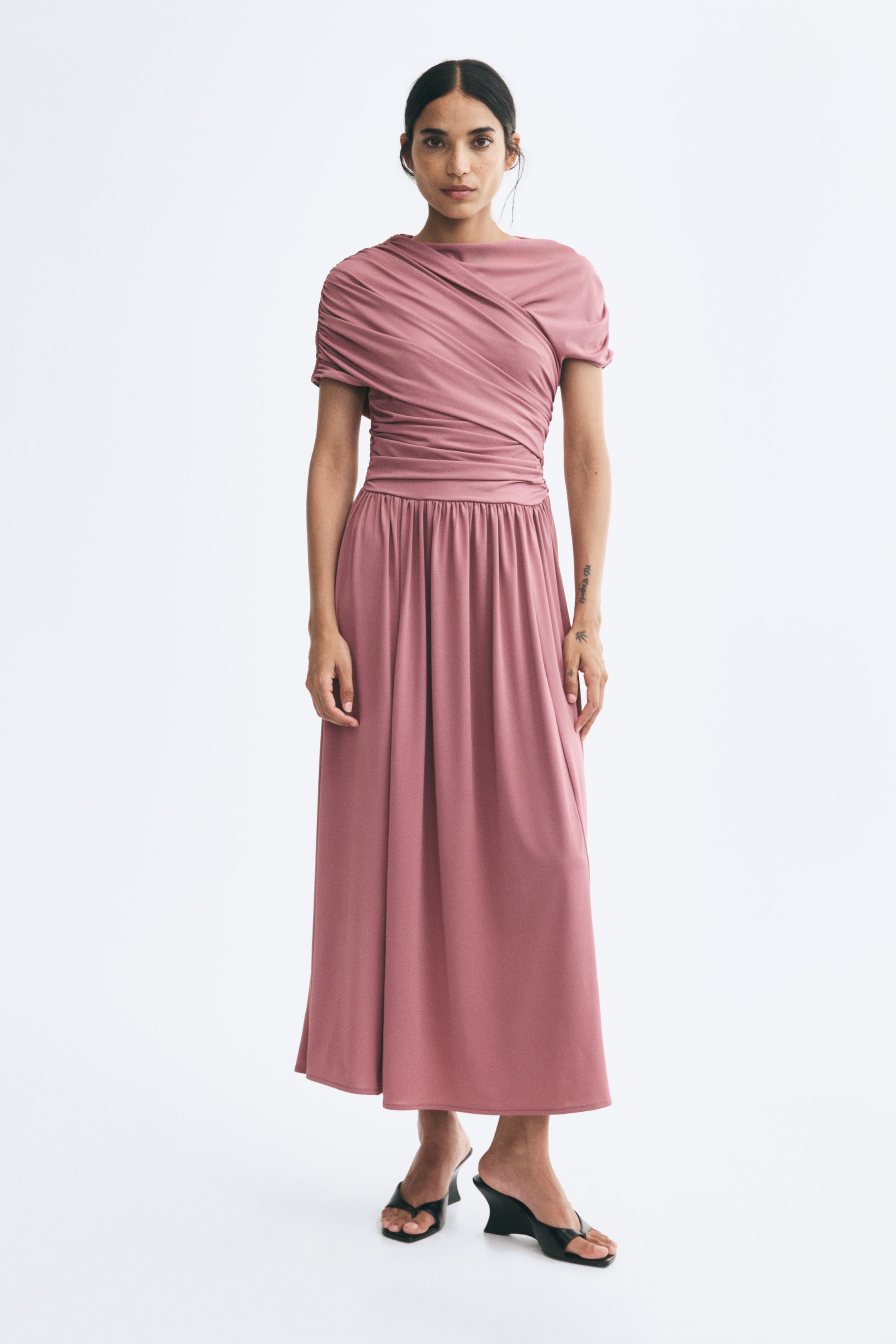 View larger image: A pink, long stretch-jersey dress is worn by the model, featuring a draped, asymmetrical bodice with gathered detailing and a boat neck. The dress has a fitted waist and a gently flared, gathered midi-length skirt, and is styled with black open-toe wedge heels.