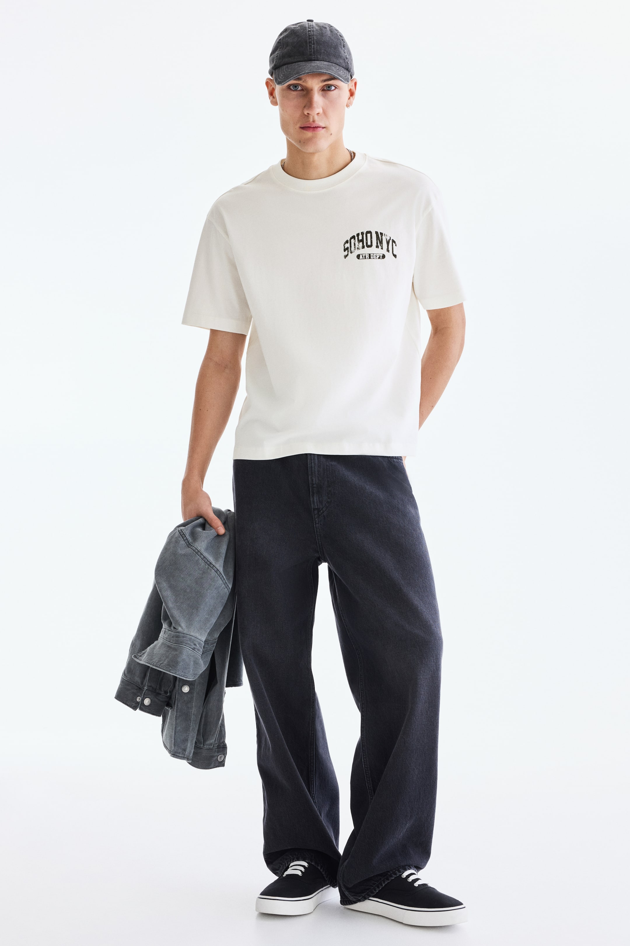 View larger image: The model is wearing a cream boxy-style t-shirt with a loose fit, featuring "SOHO NYC" print motifs. It includes a ribbed crew neck, dropped shoulders, and a straight-cut hem.