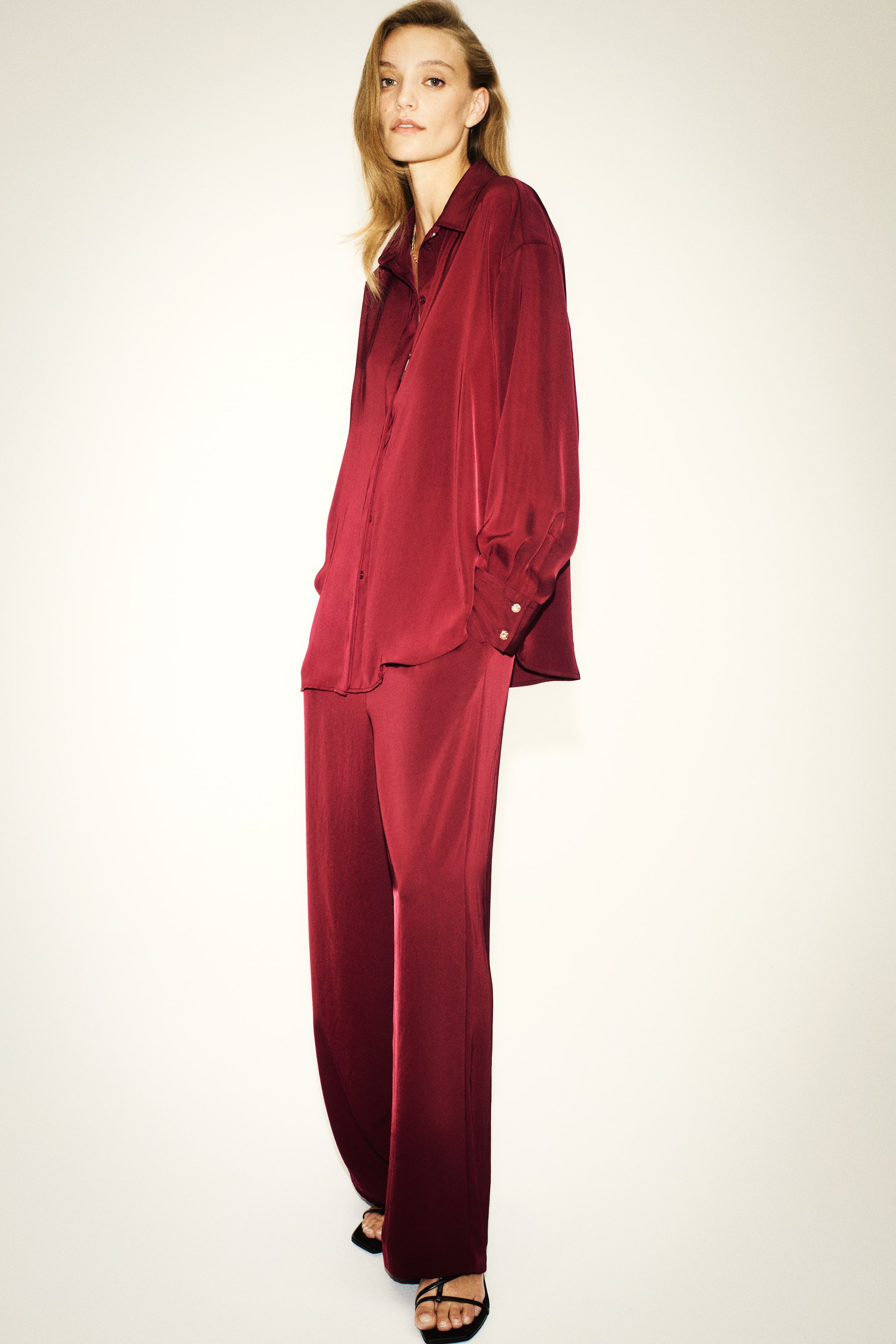 Xem ảnh lớn hơn: Chiếc quần ống thẳng bằng vải satin twill màu đỏ burgundy này có phom dáng suông rộng, mềm rủ, được chụp nghiêng sang phải để làm nổi bật độ bóng sang trọng và phom dáng thư thái. Sản phẩm được phối cùng áo sơ mi satin tay dài đồng bộ, nhấn mạnh độ rũ thanh lịch của chất vải.