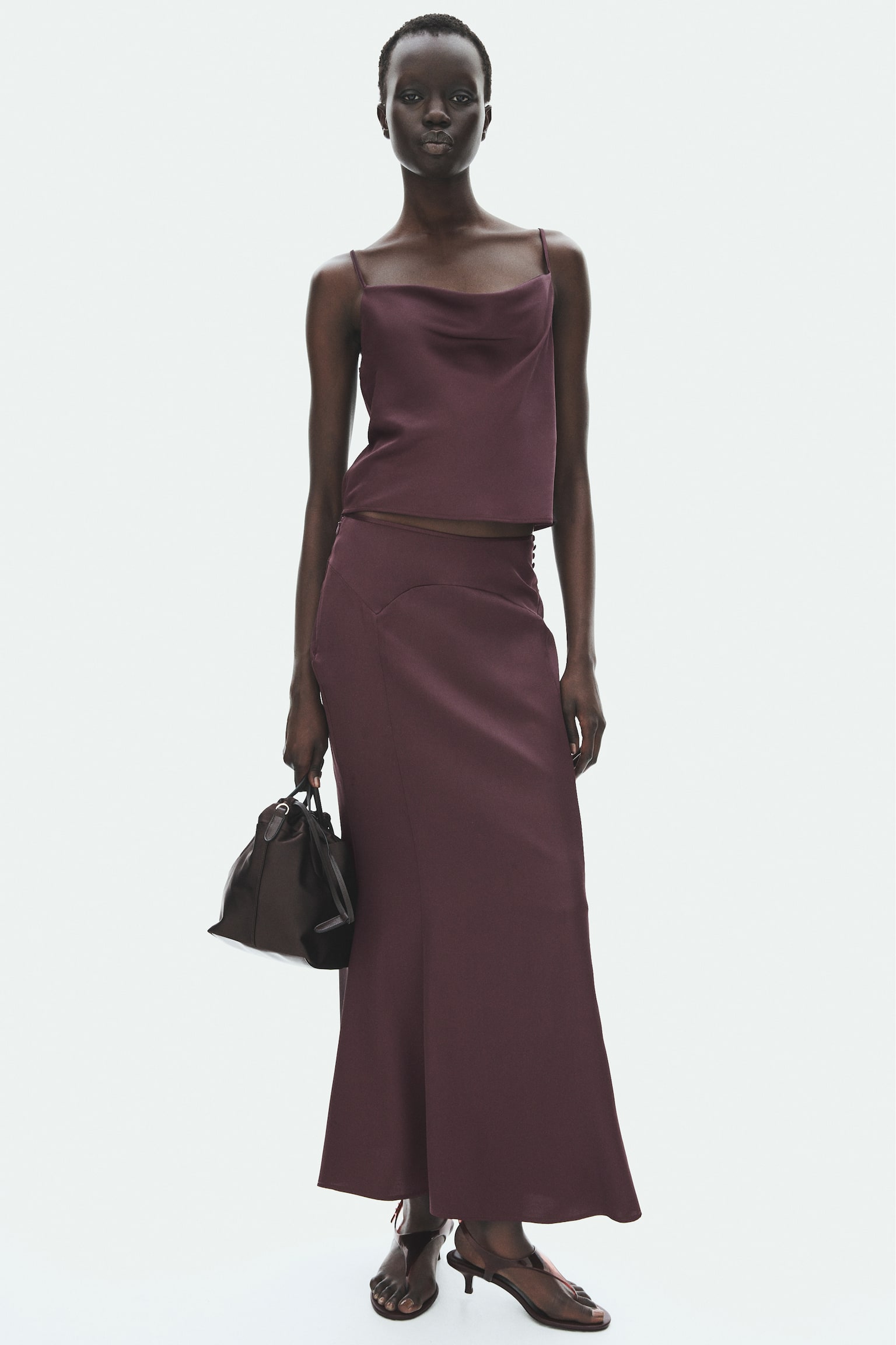 A dark plum satin-like midi skirt showcases a curved yoke detail at the waist. A row of small fabric-covered buttons runs down its right side, complementing its gently flared A-line silhouette. - 1