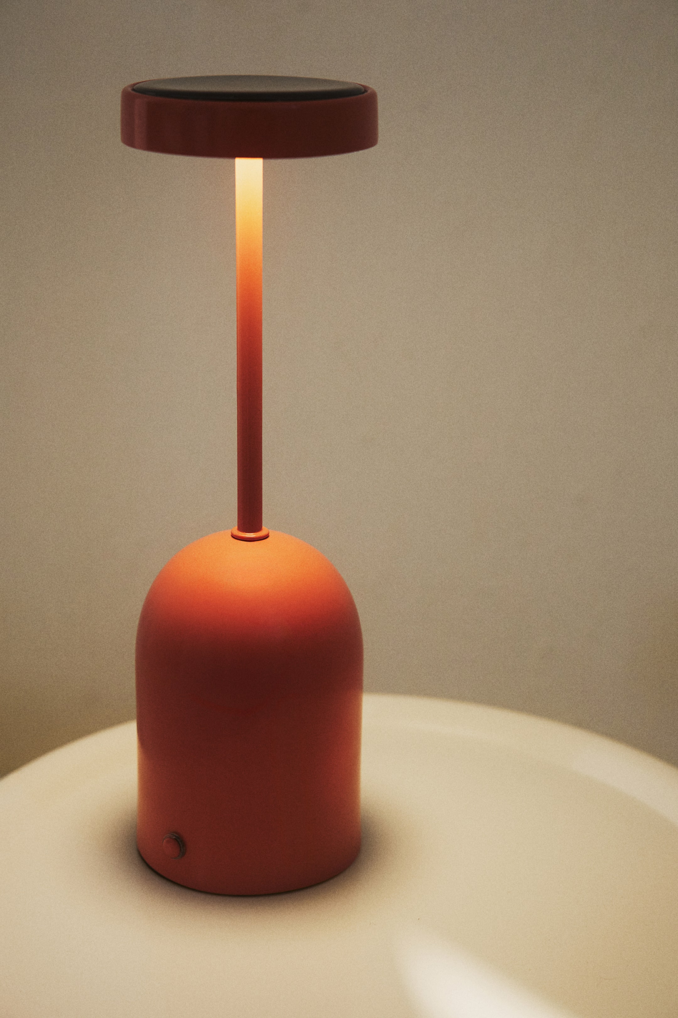 Größeres Bild ansehen: Eine korallorange pulverbeschichtete Metall-Solartischlampe präsentiert sich von vorne und besticht durch ihr schlankes Design. Ein dunkles, rundes Solarpanel krönt die Lampe, und warmes Licht strahlt nach unten von ihrem schlanken Stiel, der mit einem abgerundeten, glockenförmigen Sockel verbunden ist, an dessen unterer Vorderseite ein sichtbarer Ein-/Ausschalter angebracht ist.