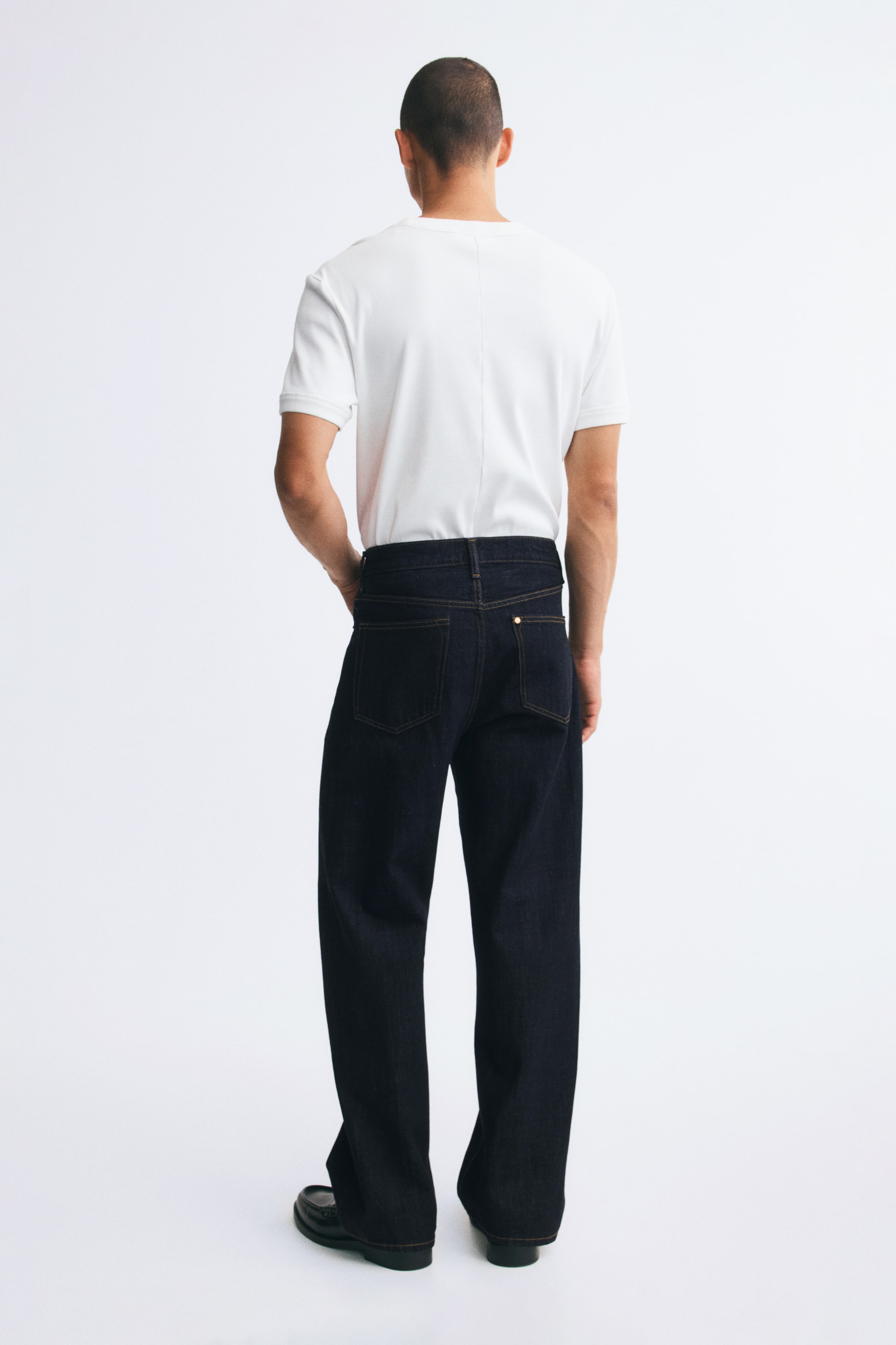 Visualizza immagine più grande: Jeans in denim di colore indaco scuro, caratterizzati da una vestibilità ampia e rilassata e da tasche posteriori applicate con cuciture a contrasto, indossati dalla modella in piedi e di spalle rispetto alla fotocamera. I jeans a vita regolare sono abbinati a una t-shirt bianca infilata all’interno e a scarpe nere.