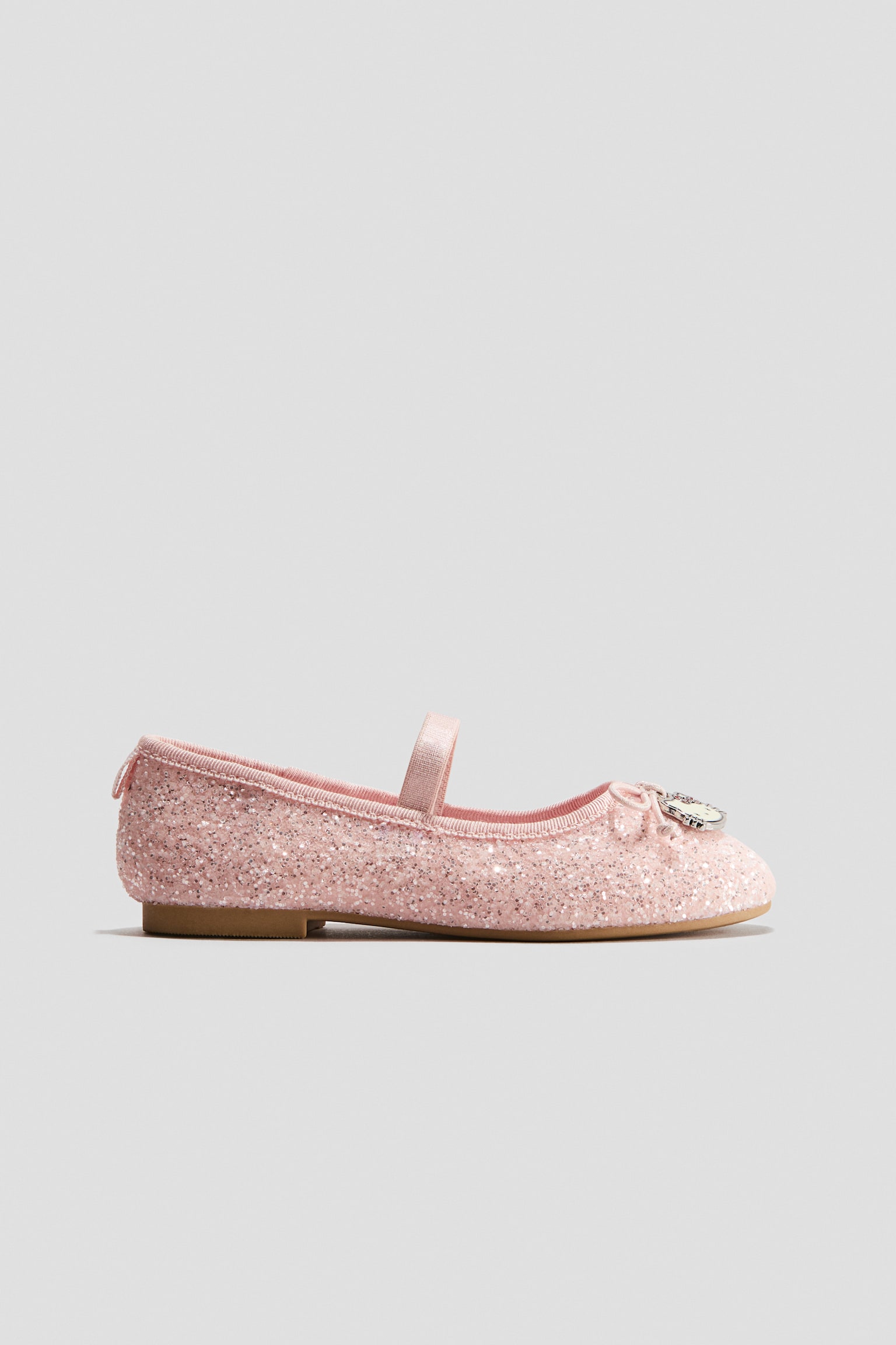 A pink glittery ballet flat is shown in a side profile facing right, featuring a matching pink elastic strap across the instep and a grosgrain trim around the opening. The shoe is designed with a rounded toe adorned with a decorative bow and a small metal charm, finished with a pull loop at the heel and a flat tan sole.