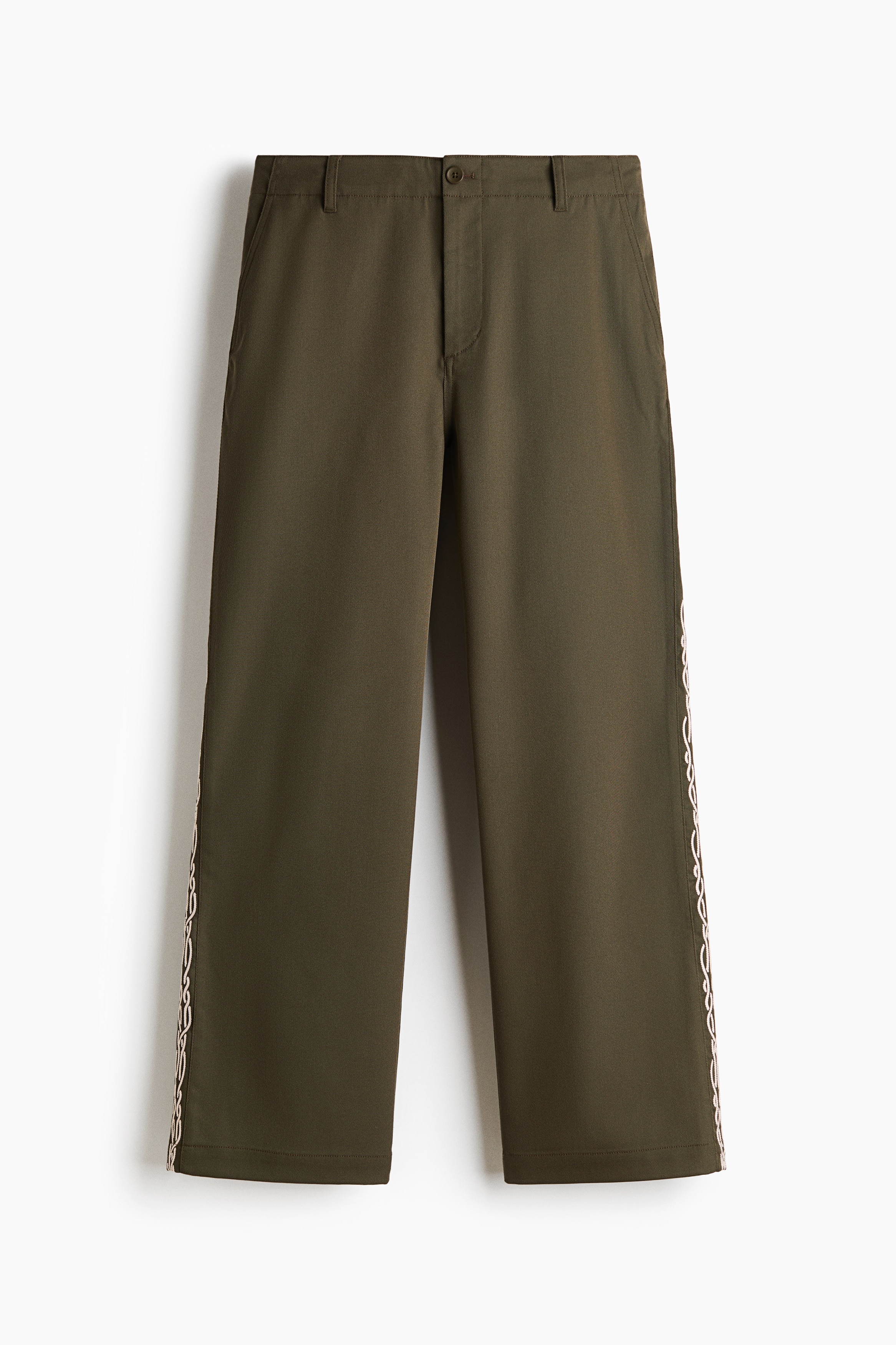 The model is wearing olive-green relaxed-fit chinos featuring a cream-coloured embroidered chain pattern running down the side of the leg. The trousers are styled with a black belt and a white tank top, while a matching olive-green jacket is being held in one hand.