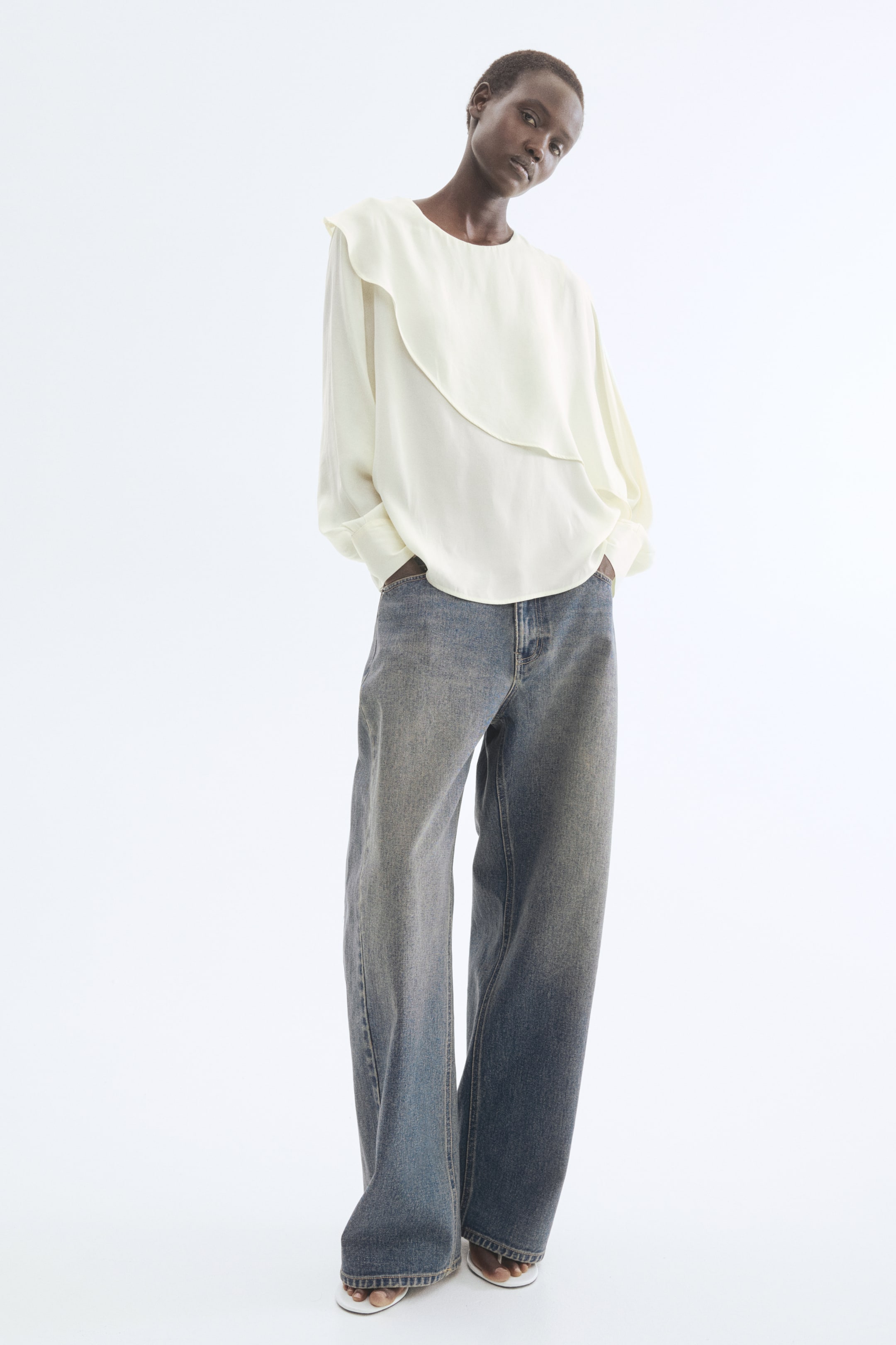 Prikaži veću sliku: Model nosi svetlu bluzu sa okruglim izrezom, dugim rukavima i manžetama na dugmad. Bluza ima asimetričan slojeviti panel koji se spušta preko prednje strane, a model stoji sa rukama u džepovima širokih farmerki.