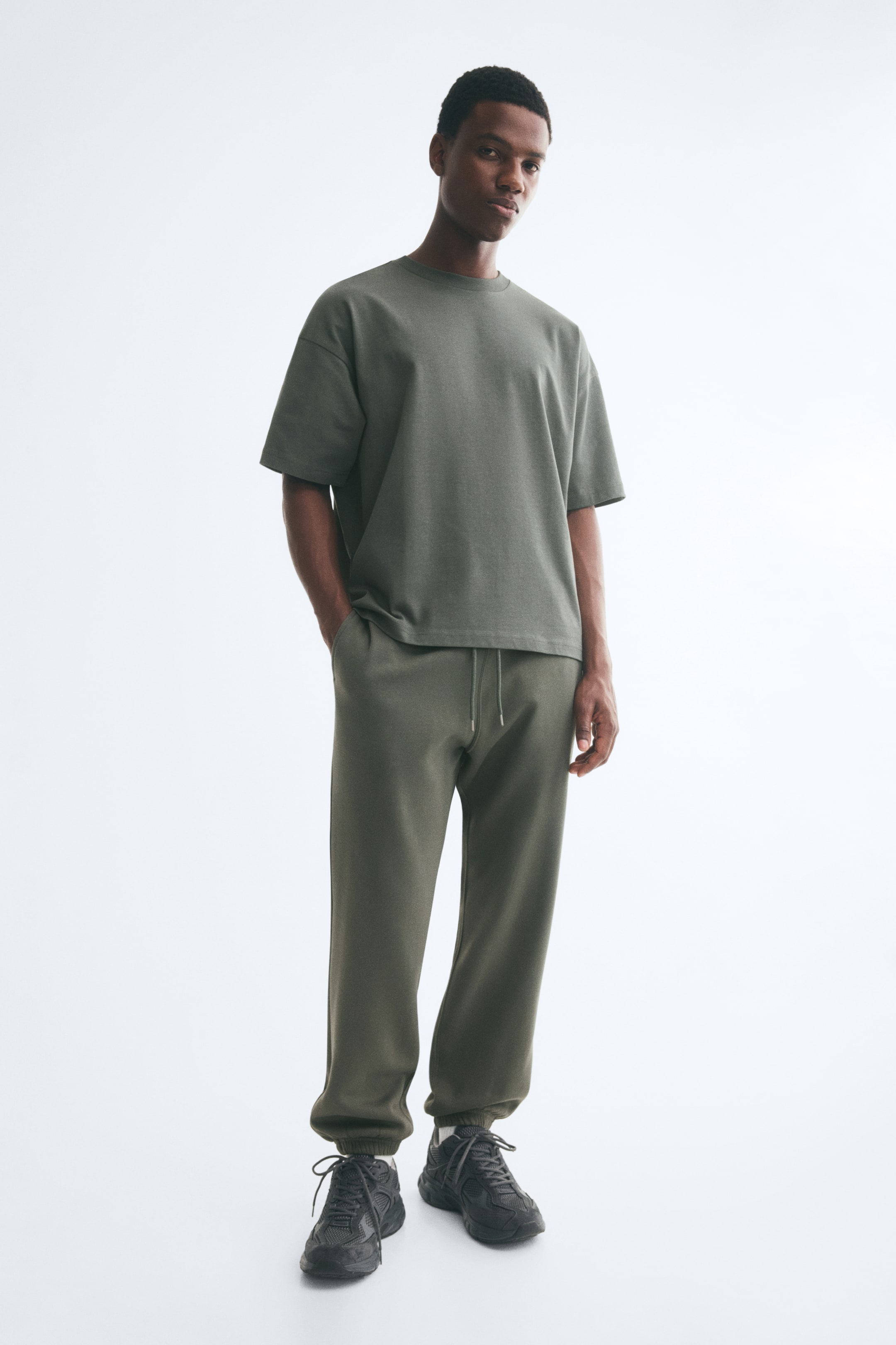 View larger image: The model is wearing olive green sweat fabric track pants, featuring an elasticated drawstring waistband, side pockets, and ribbed hems. The model is posing with one hand placed in a side pocket, styling the trousers with a matching olive green T-shirt and dark trainers.
