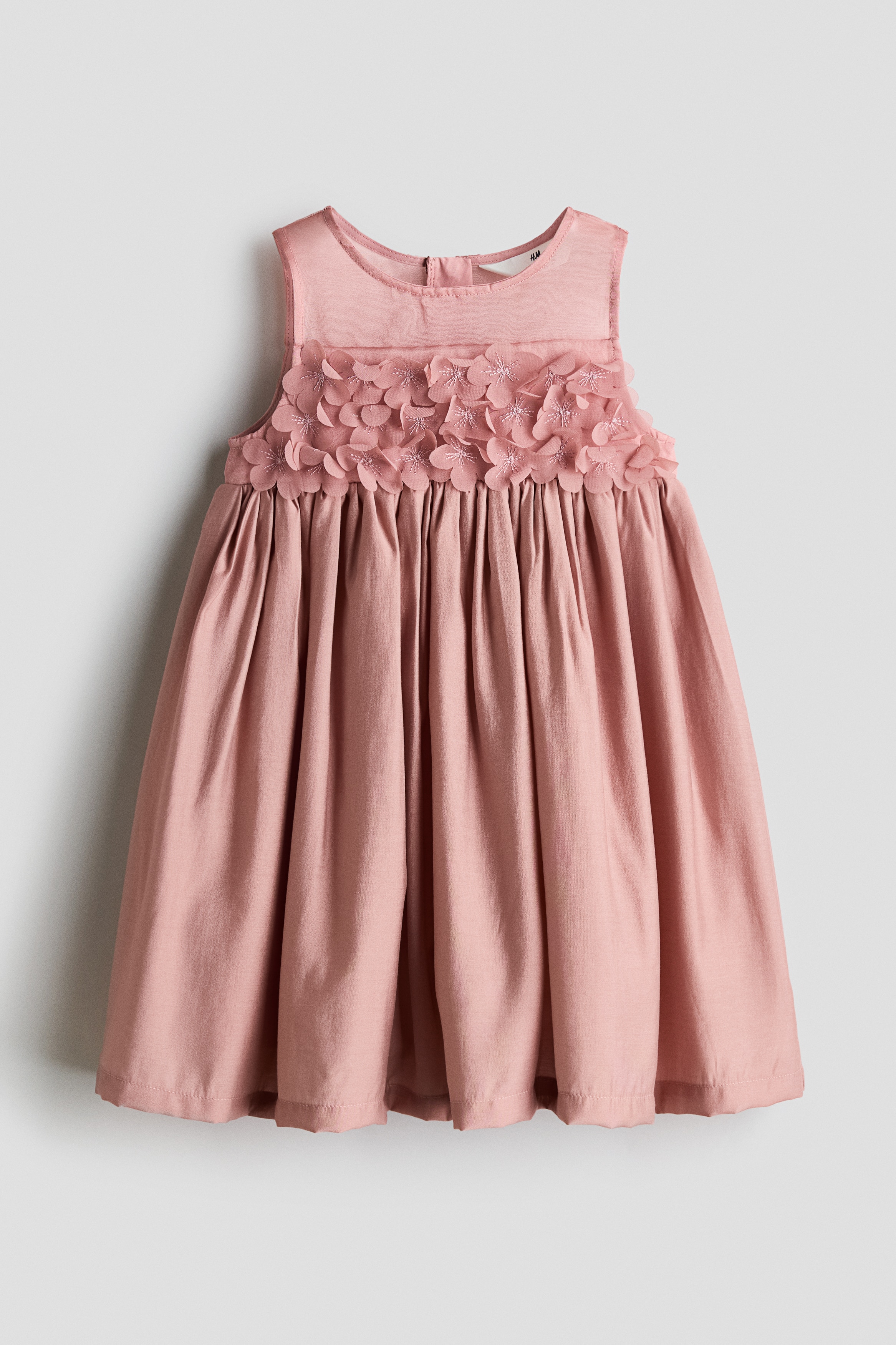 A sleeveless, dusty pink dress is featured from the front, showcasing a round neckline and three-dimensional floral appliquÃ©s across the bodice. The dress falls into a gathered, flared skirt from a defined waistline.