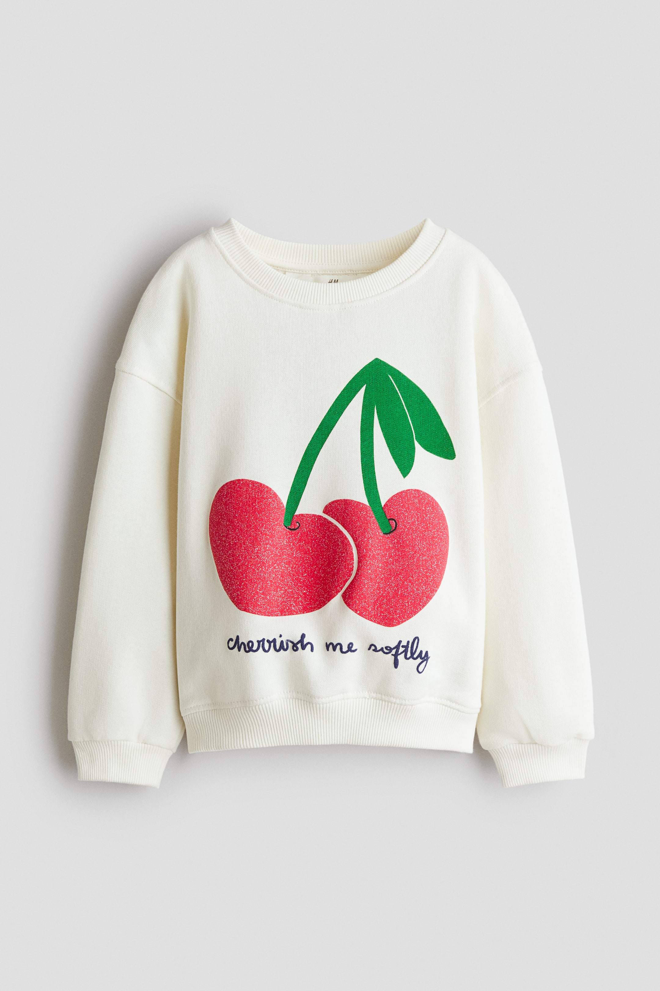 View larger image: A cream sweatshirt is shown from the front, featuring a large red cherry motif with green stems and leaves, and the text "cherish me softly" printed below it. It is designed with a round, rib-trimmed neckline, dropped shoulders, and ribbed cuffs and hem.