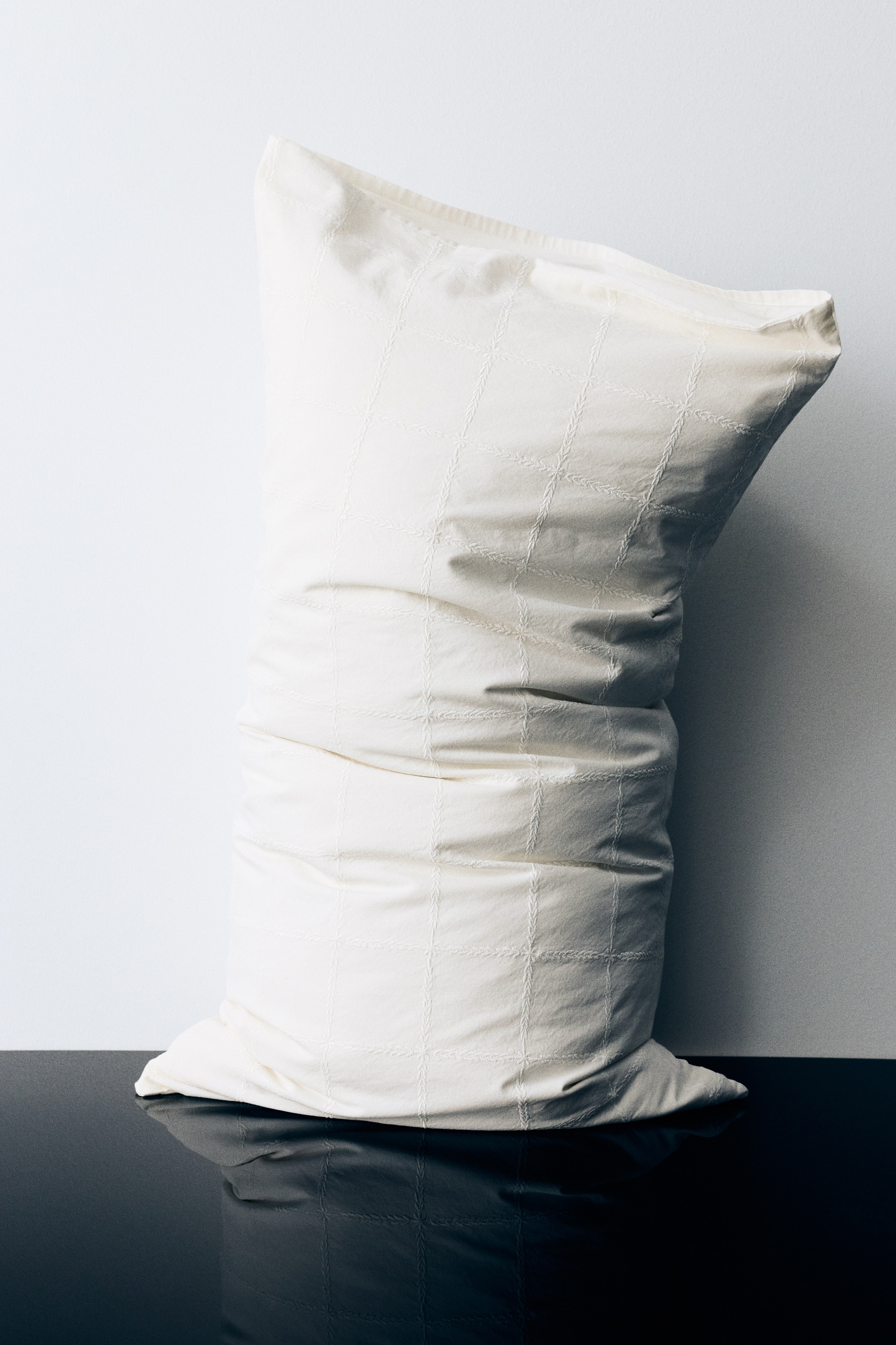 A long, rectangular, cream-coloured cushion is lying horizontally, displaying a stitched grid pattern across its slightly crinkled surface.