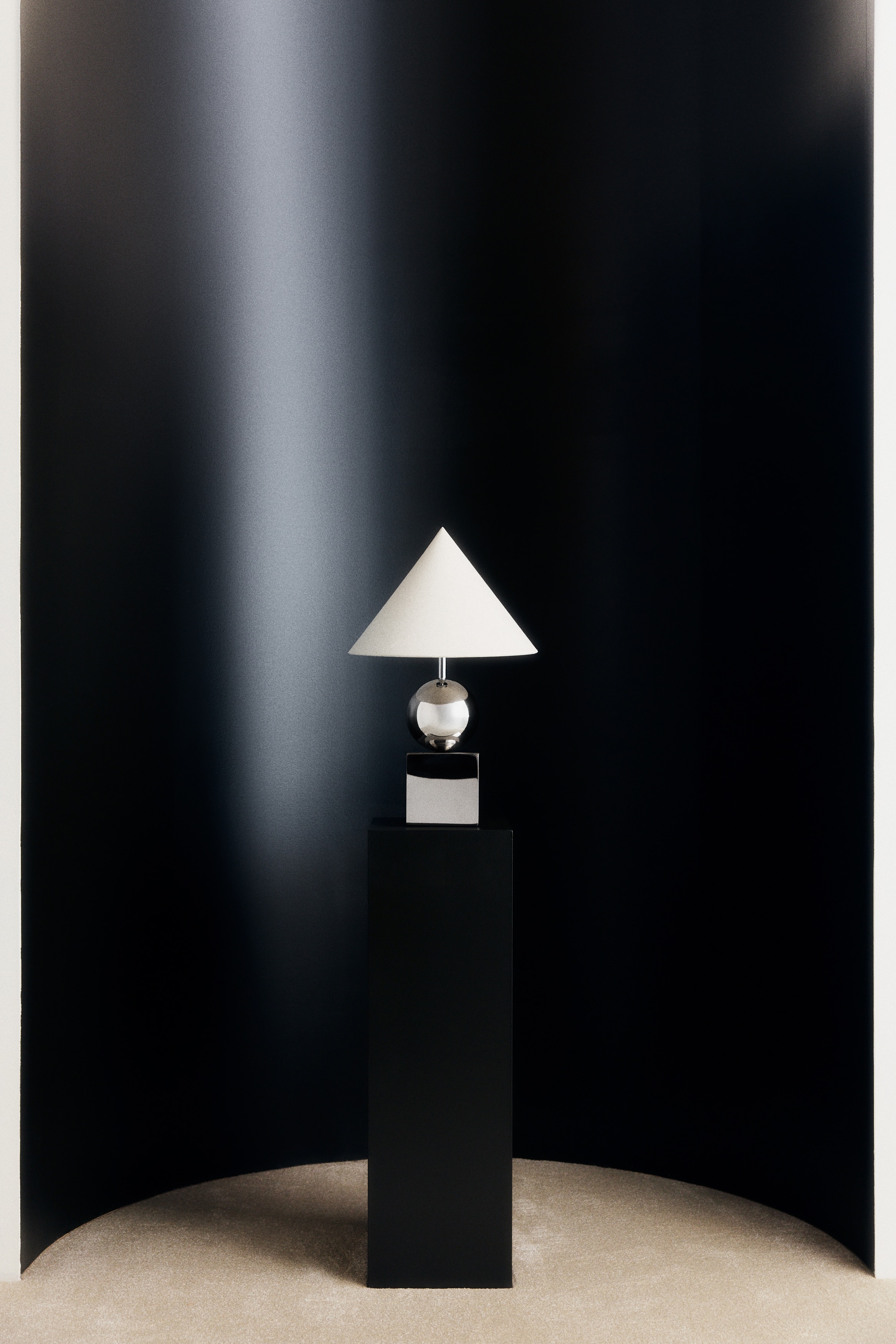 The table lamp is showcasing its front view, featuring a conical, off-white fabric shade. This shade is resting on a slender chrome stem that emerges from a polished chrome sphere, which is positioned on a square, polished chrome base with a black power cable extending from its rear.