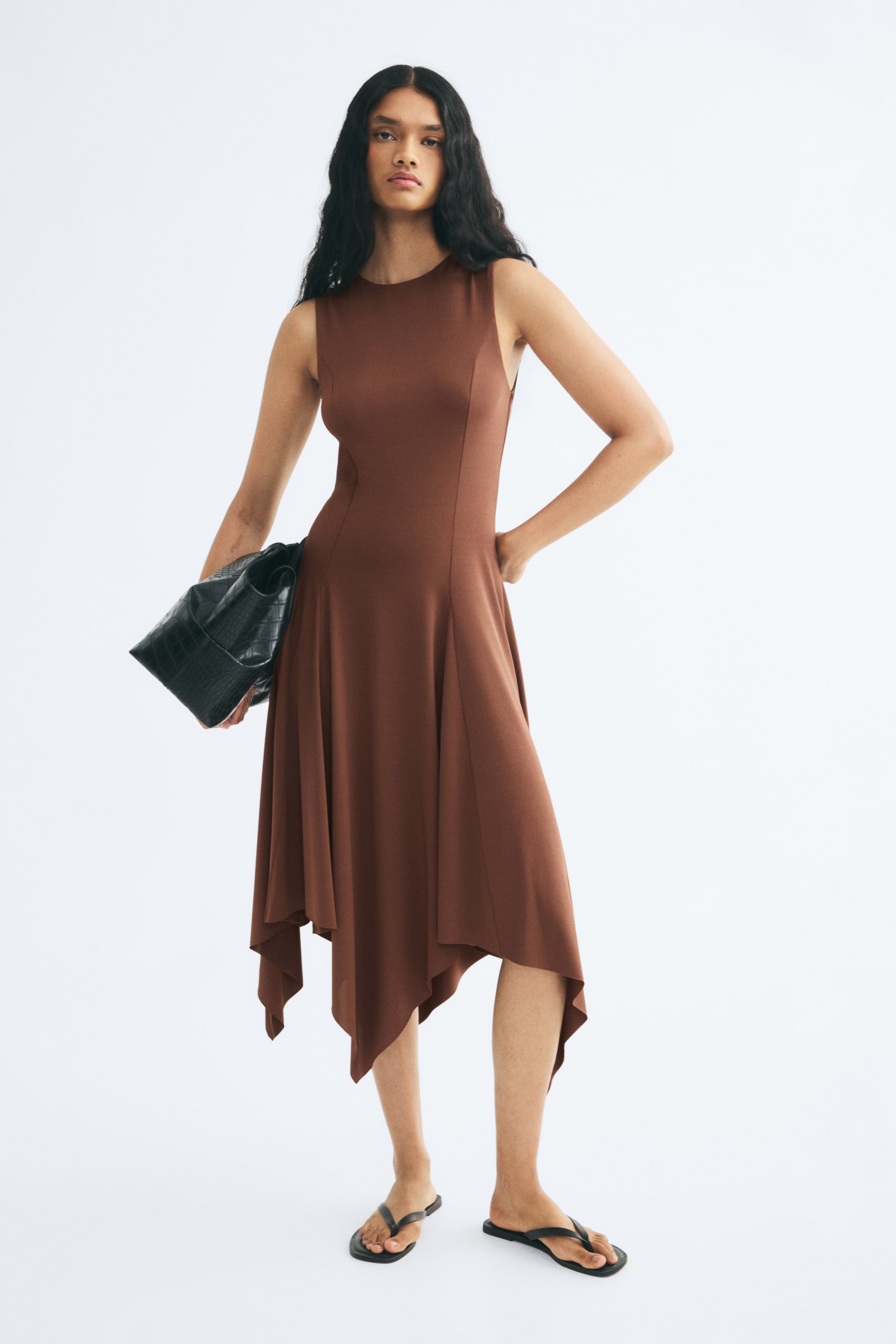 View larger image: A brown, sleeveless, slim-fit dress with a round neckline is worn by the model. The dress features princess seams that open into a flared skirt with godet pleats and an asymmetrical handkerchief hem, styled with a black handbag tucked under the arm and black flat sandals.