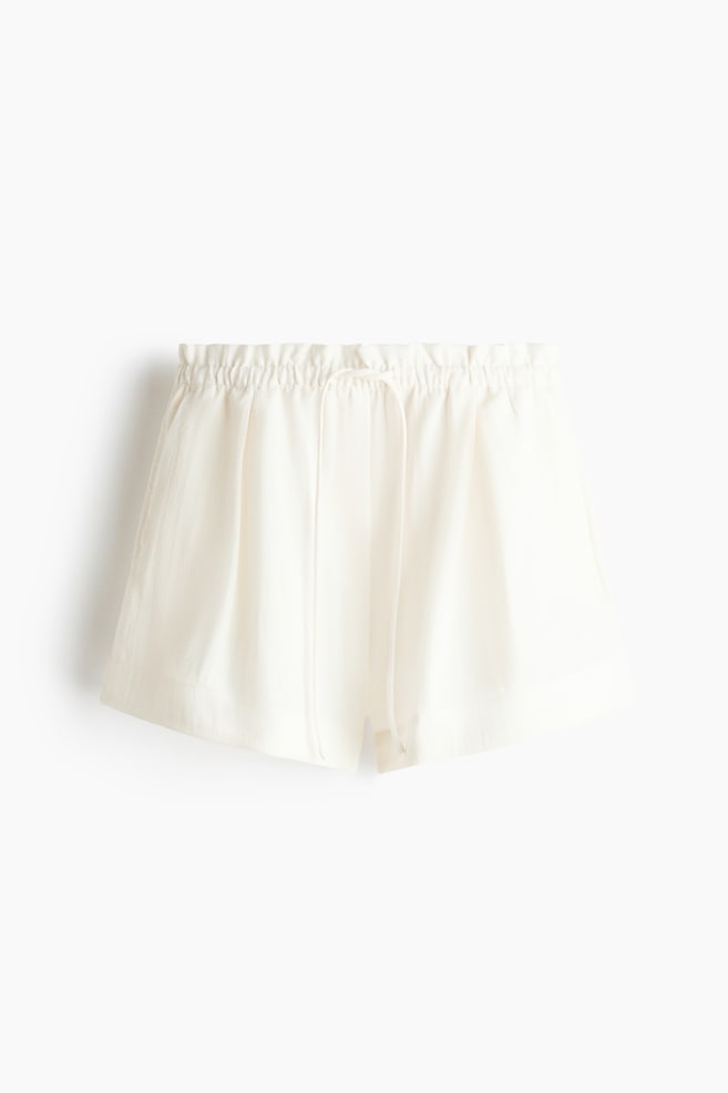 The cream-coloured shorts feature an elasticated, gathered waistband with a drawstring tie at the front. The loose-fitting shorts showcase small side slits at the hem. - 1