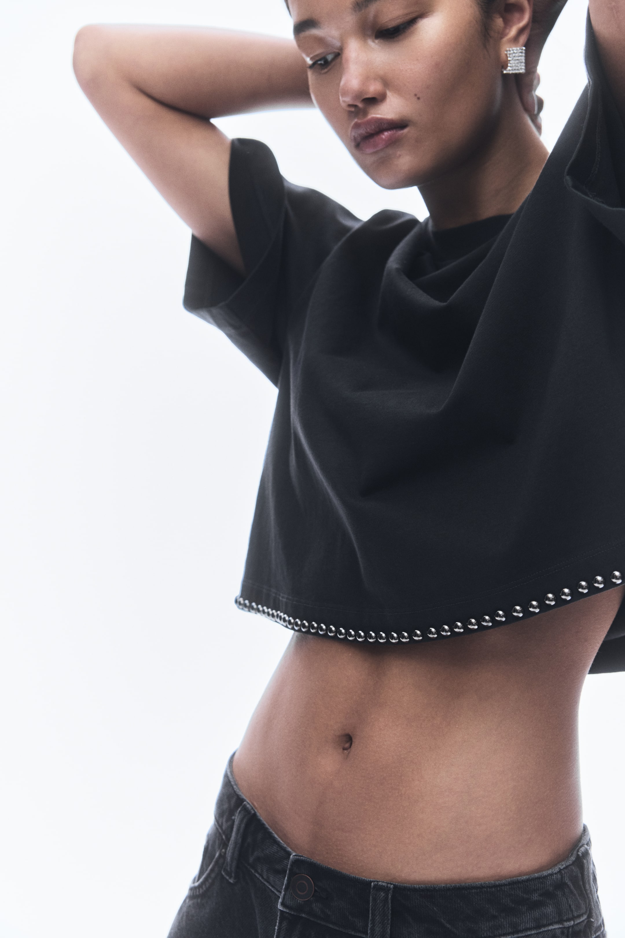 View larger image: A black short-sleeved cropped top is being modeled, featuring a relaxed fit and a hemline intricately embellished with a continuous row of small, silver-toned studs.