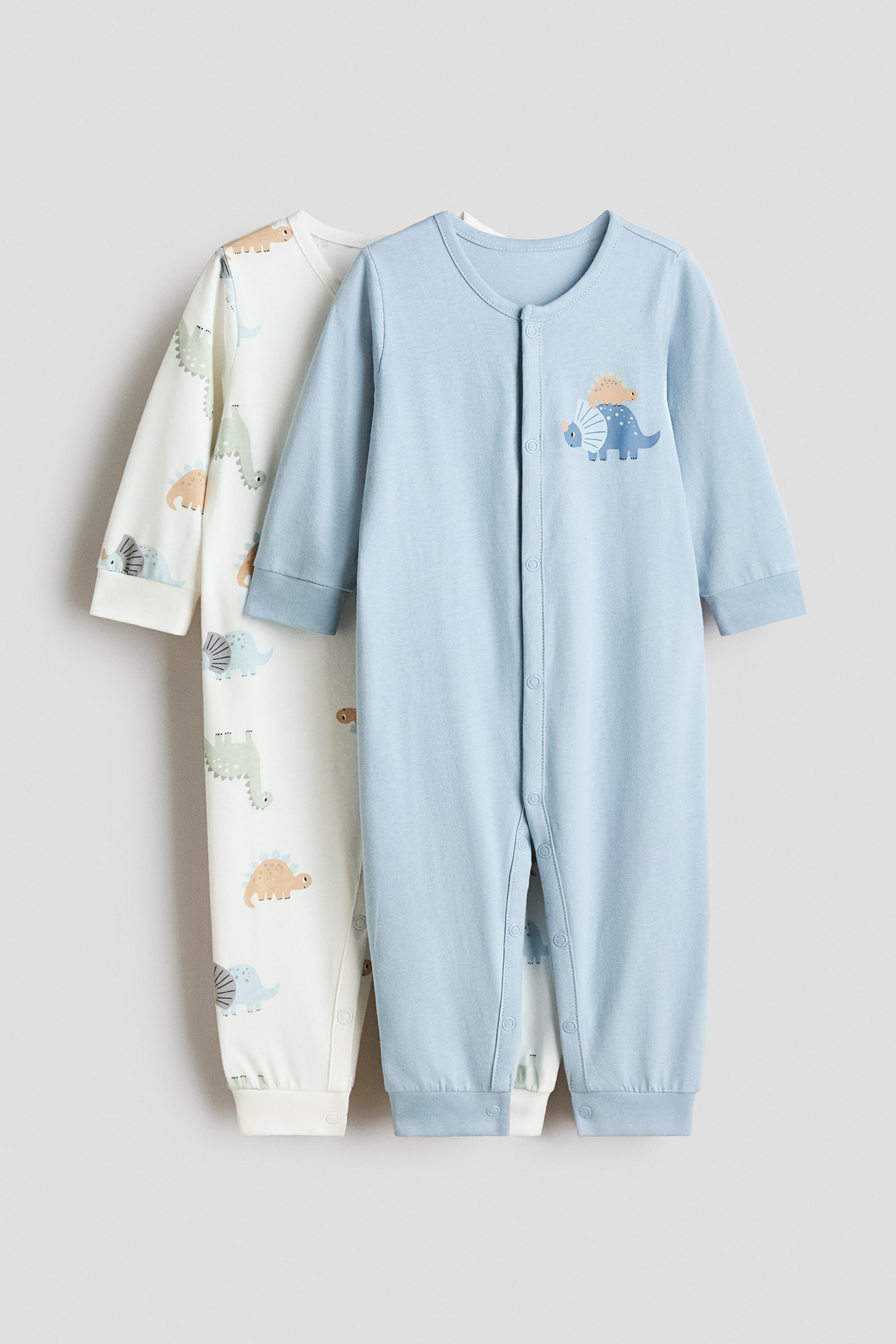 Xem ảnh lớn hơn: Hai bộ đồ ngủ làm bằng vải jersey siêu mềm đang treo lên, cả hai đều có tay dài và viền cổ tay cùng gấu quần có gân. Bộ đồ ngủ màu xanh nhạt có cúc bấm trải dài từ trước ra chân, với hình ảnh khủng long trên ngực. Bộ đồ ngủ màu trắng ngà có in hình khủng long toàn bộ và cùng kiểu đóng cúc bấm.