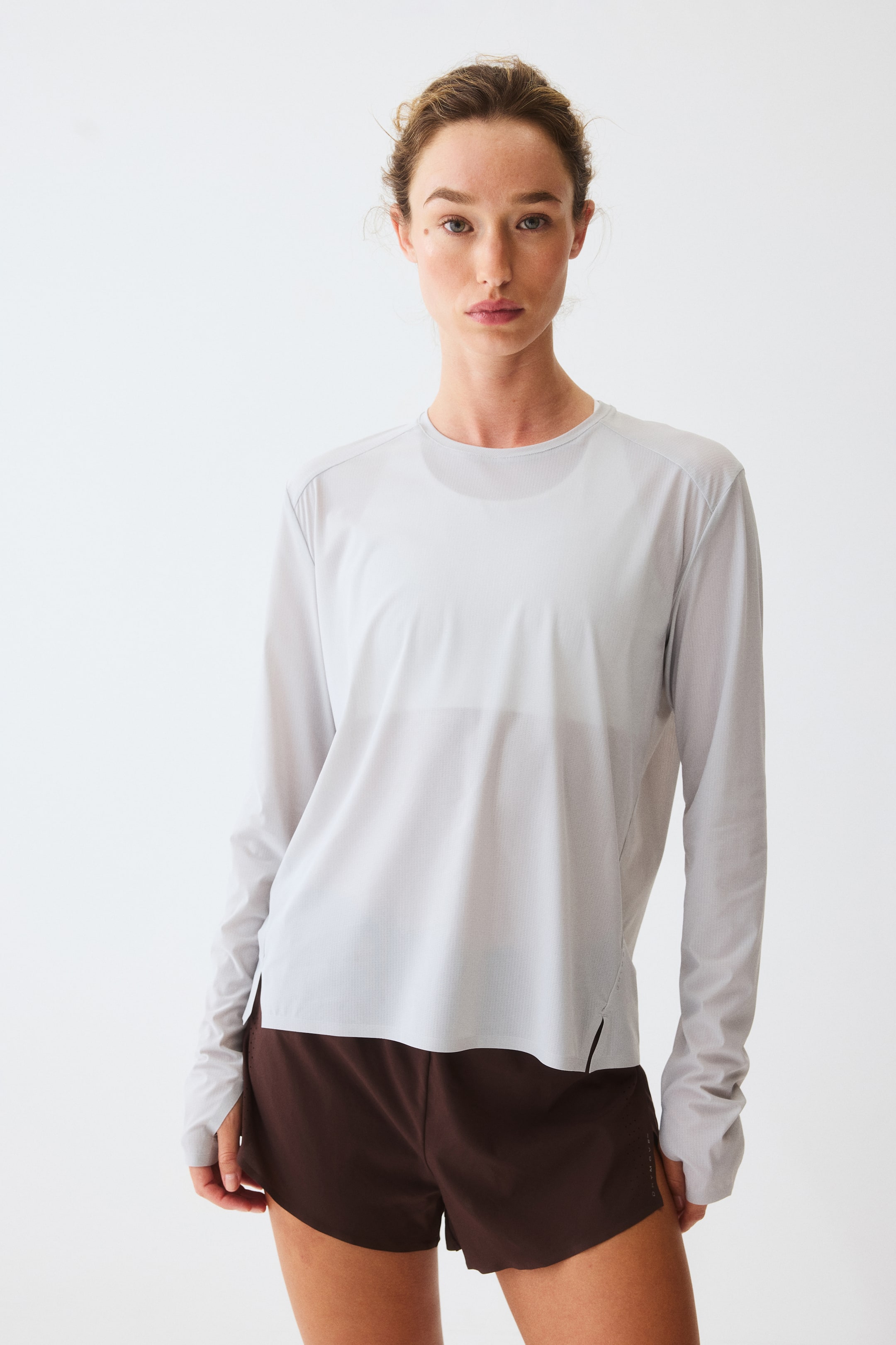 Ver imagen más grande: Se presenta una camiseta deportiva de manga larga en gris claro con un corte holgado, que cuenta con un cuello redondo y mangas raglán. La tela ligera y texturizada muestra discretas aberturas laterales en el dobladillo y una prenda más clara debajo.