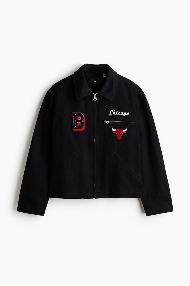 A black jacket is presenting front-facing, featuring a standard collar and a full-zip closure. It is displaying a textured red and black 'BULLS' patch on the left chest, white 'Chicago' embroidery above a zippered pocket on the right, a red, black, and white Chicago Bulls logo patch below it, and two angled welt pockets on the lower front. - 1