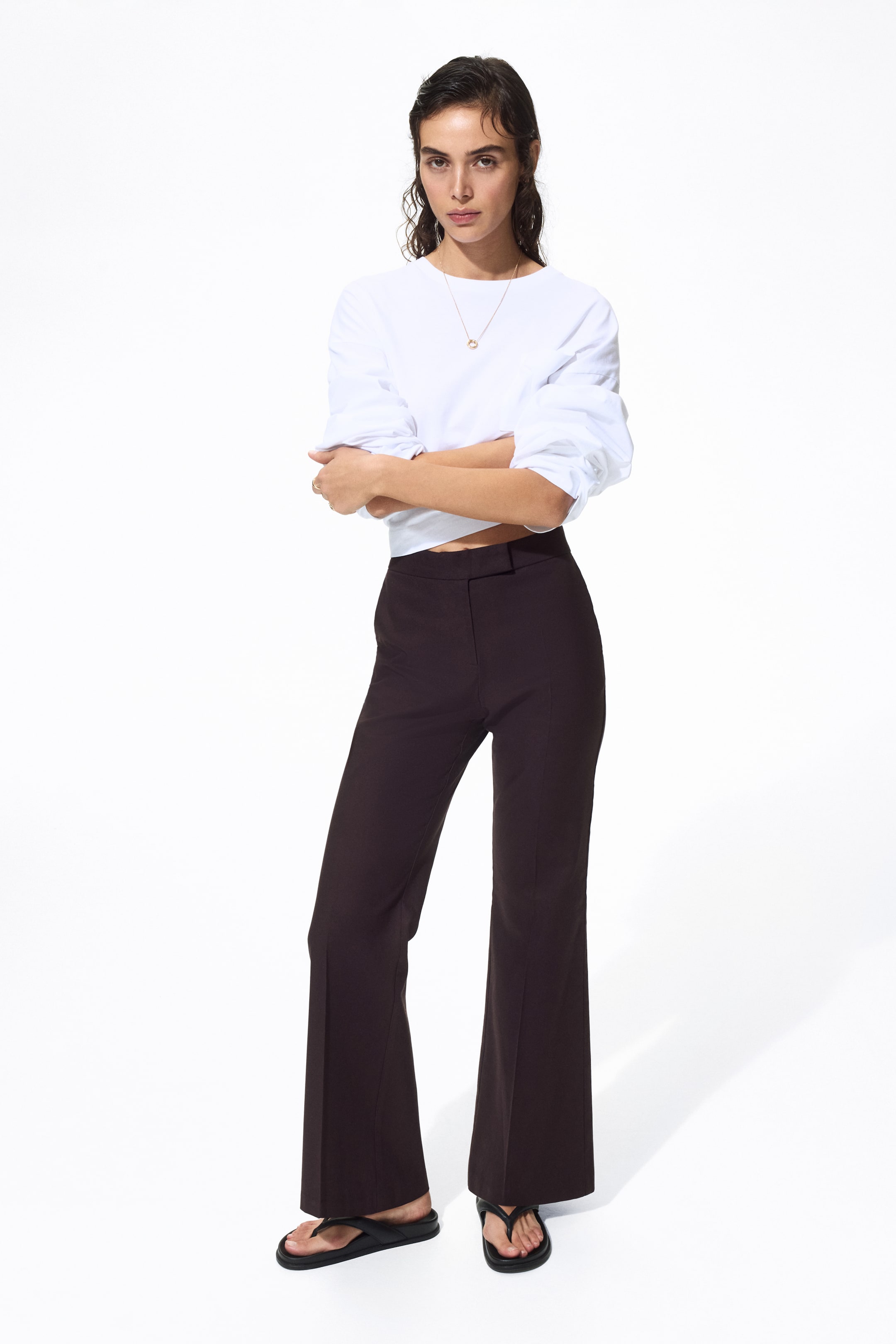 View larger image: Dark brown trousers boast a high-waisted fit and a bootcut silhouette, elegantly flaring from the knee. They showcase a tailored flat front with a vertical seam down the center of each leg, paired with a white top, and gracefully extending to the floor over black sandals.