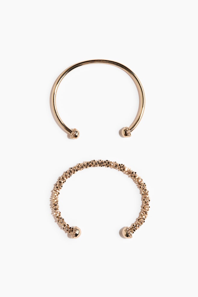 Two open-cuff bracelets in a gold tone are shown from a top-down view. The upper bracelet is featuring a smooth band with small, textured spherical ends, while the lower bracelet is showcasing a granular, textured band with larger, textured spherical ends. - 1